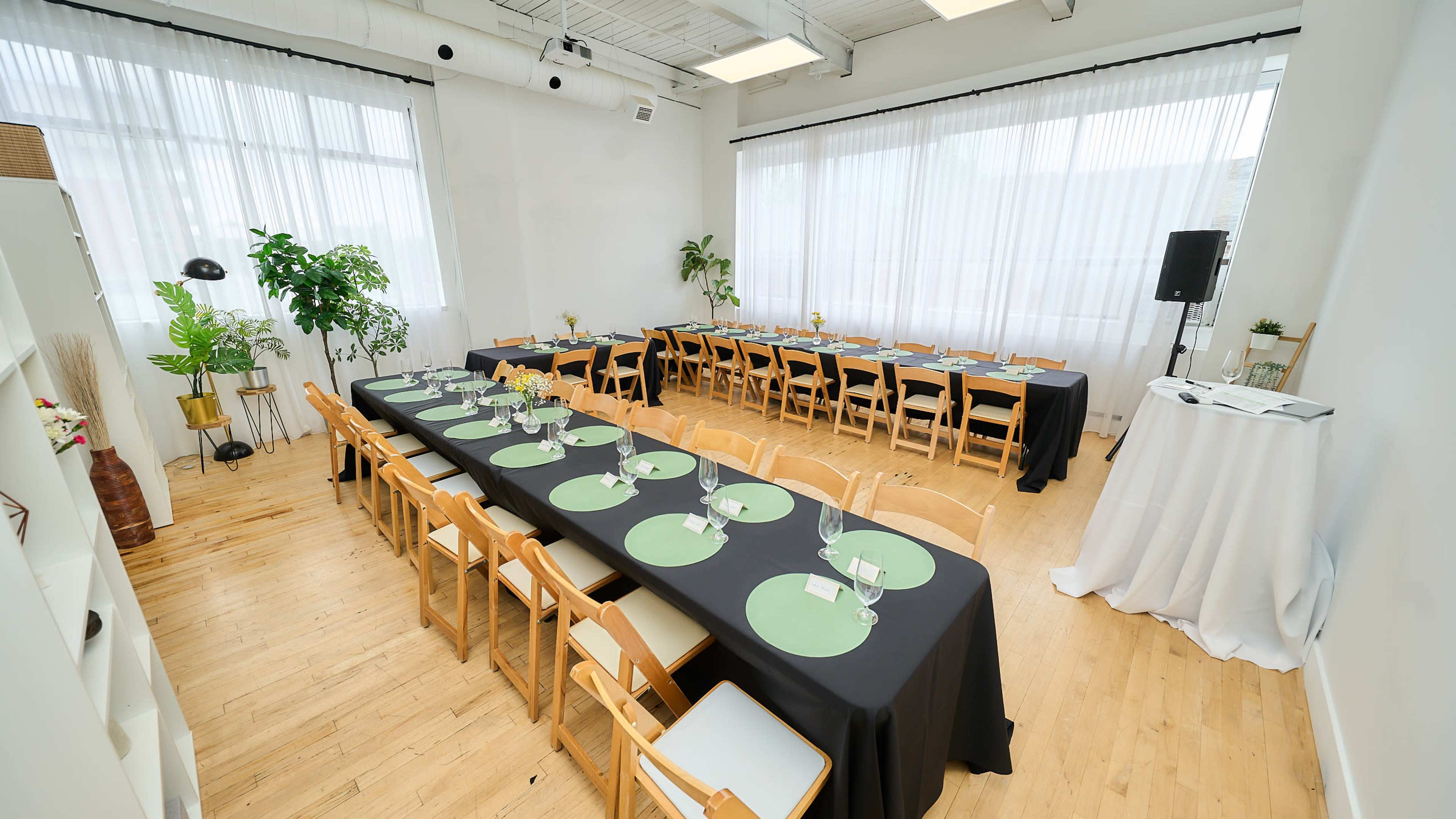 A spacious dining room features two long tables set with green placemats and chairs, surrounded by plants and natural light from large windows.