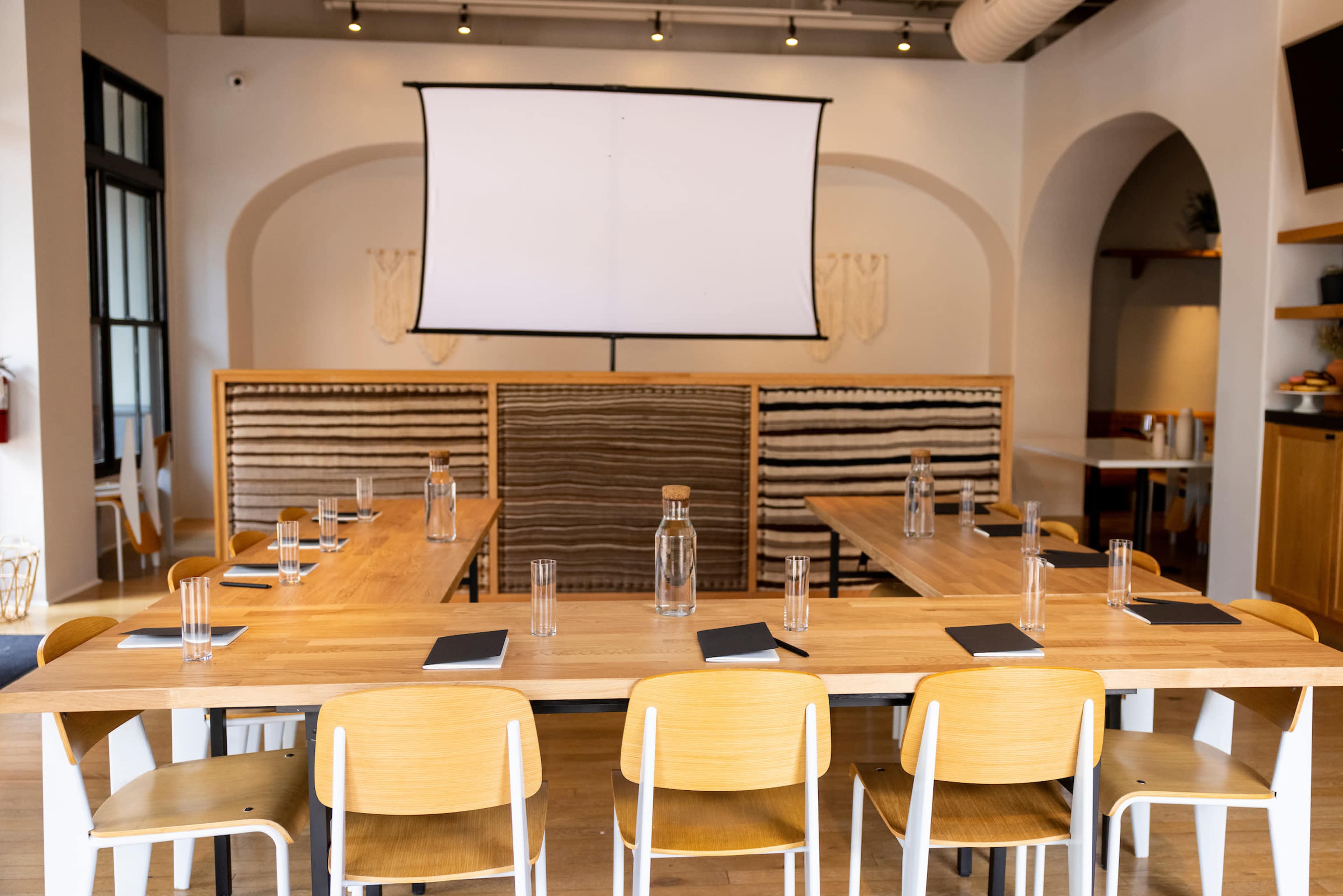 A spacious meeting room features two large rectangular tables arranged in an "H" shape, surrounded by chairs, with a blank projector screen in the center.