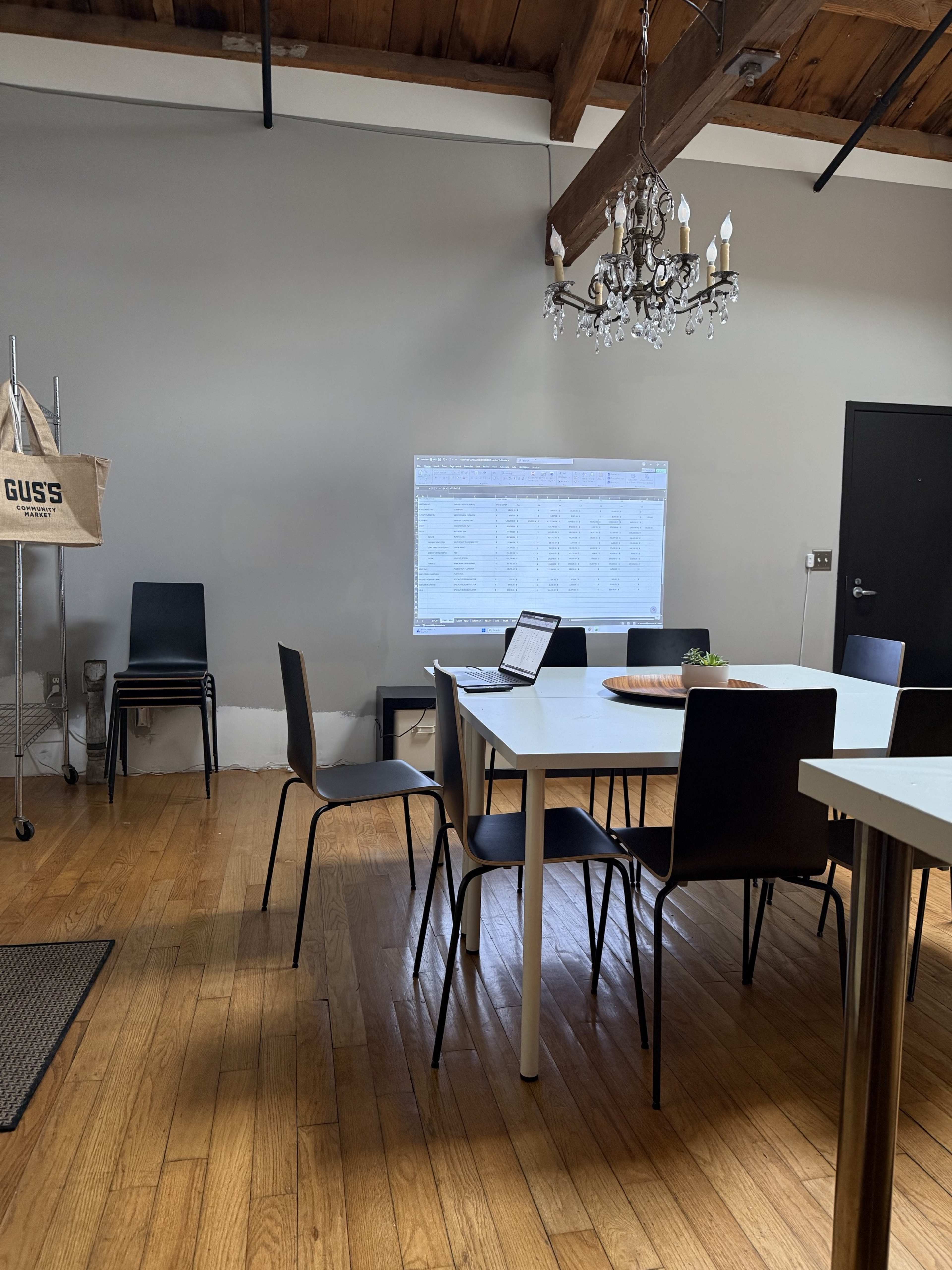 A conference room features a long white table surrounded by black chairs, with a laptop opened and a projection of a spreadsheet on the wall.