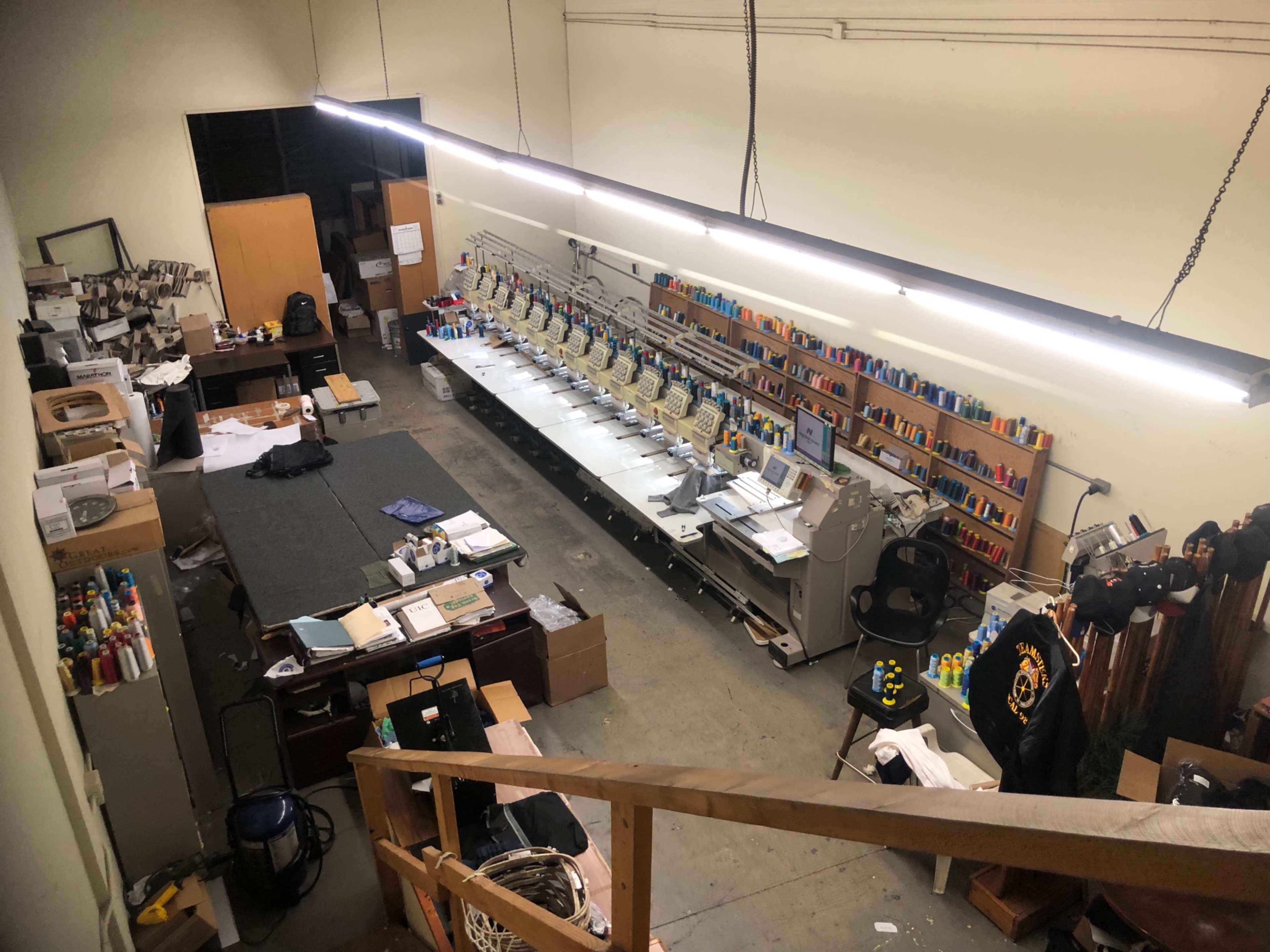 A spacious workshop with a row of sewing machines, various colorful spools of thread on shelves, and scattered materials and equipment around the floor.