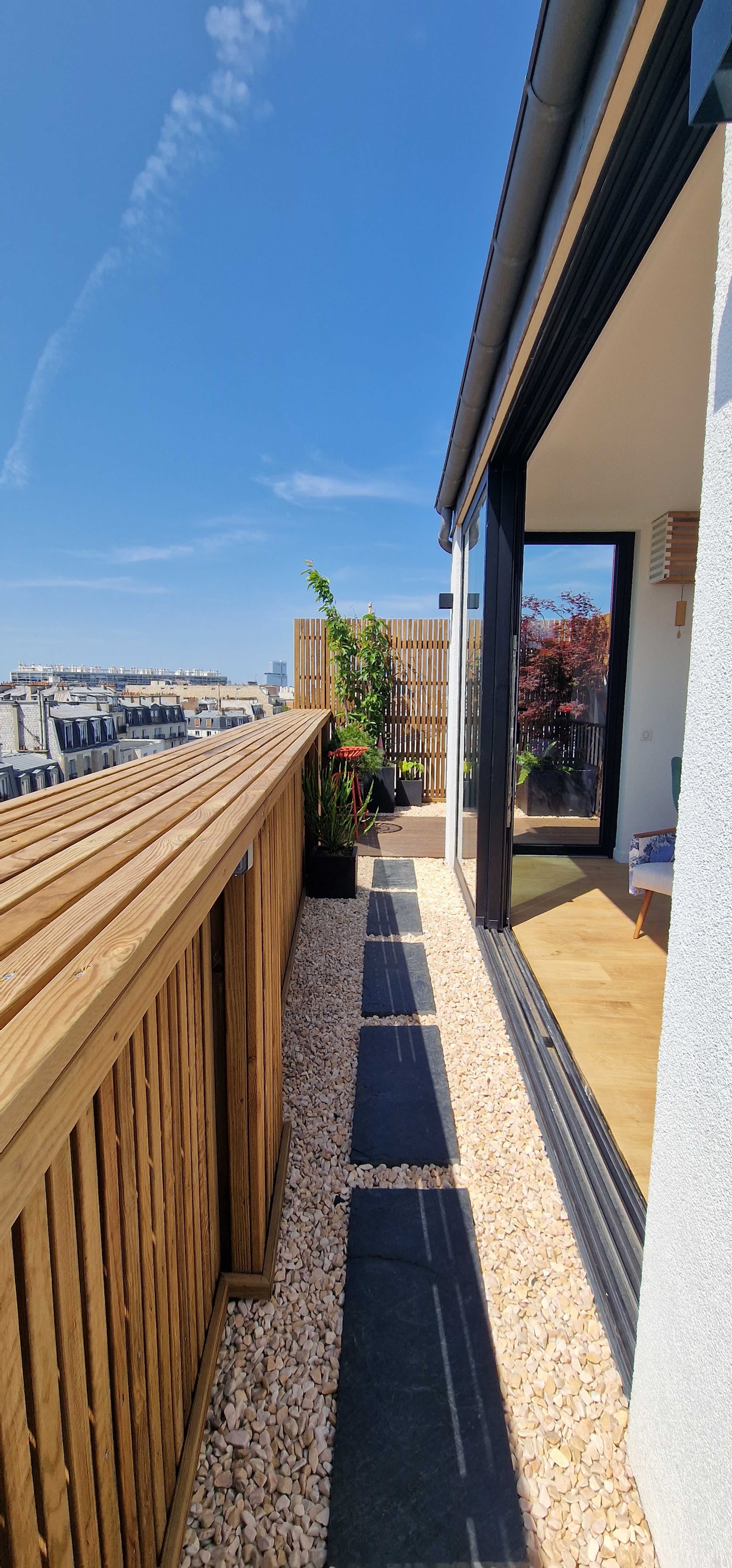 A rooftop terrace with wooden railing, stone pathways, and potted plants under a clear blue sky.