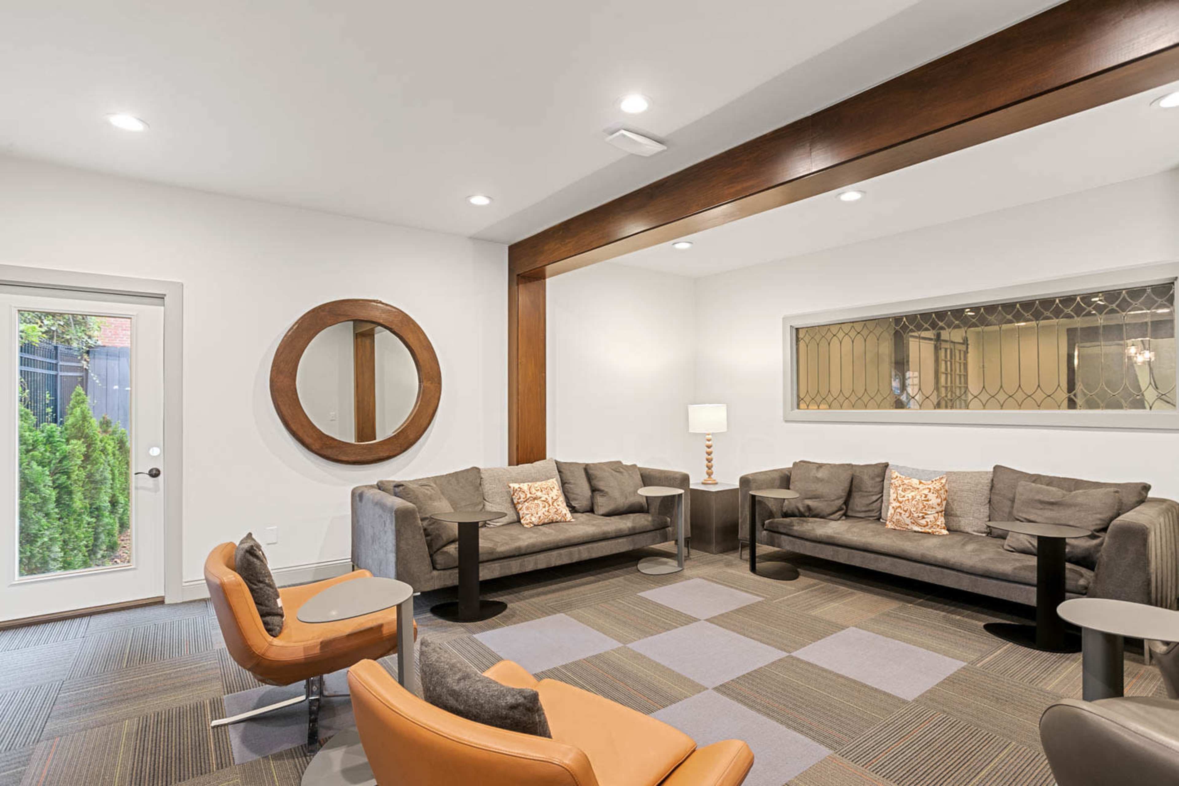 The image shows a modern lounge area featuring two gray sofas, orange chairs, and a round mirror on the wall, with a sliding glass door leading outside.
