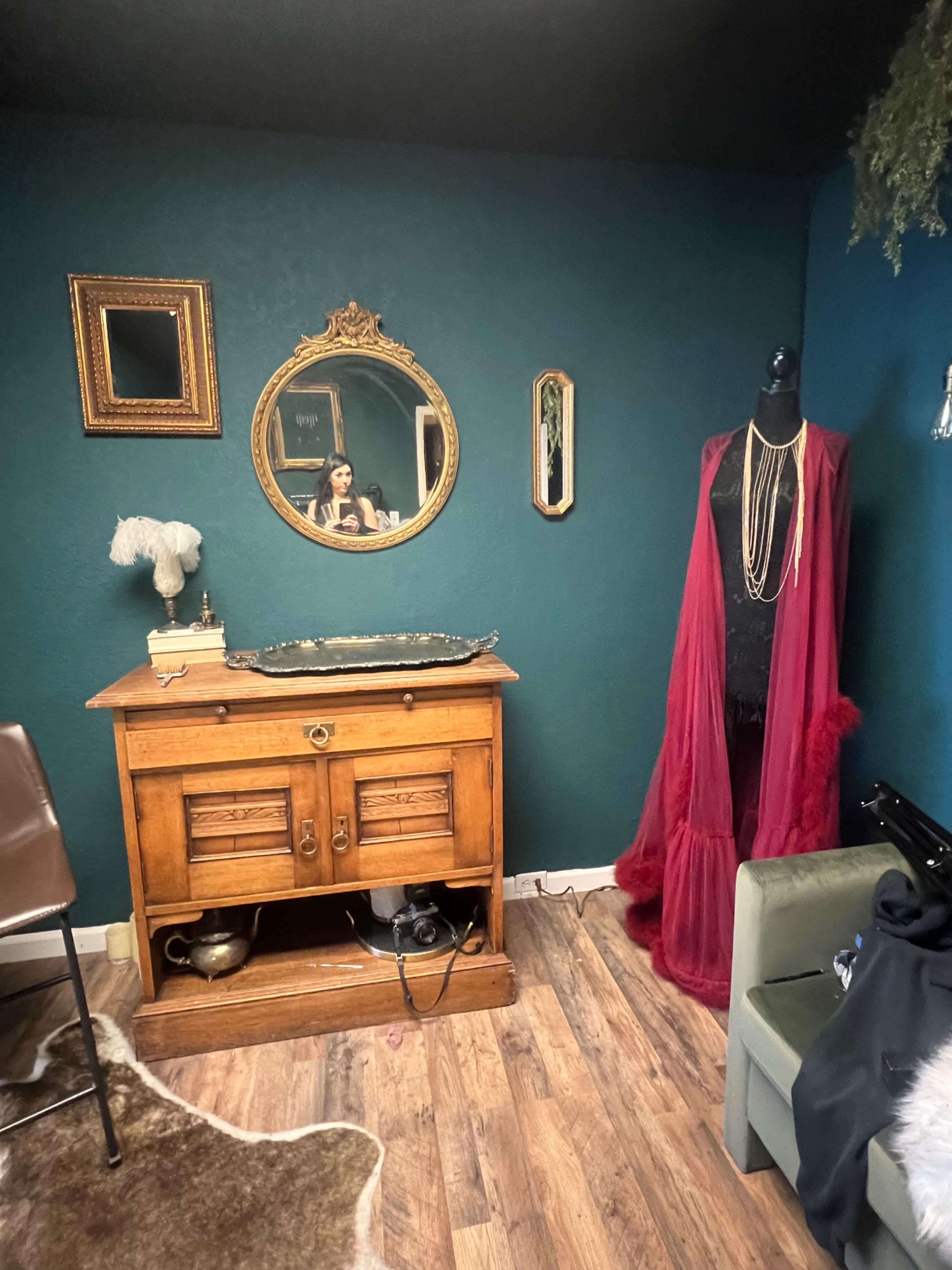 A vintage wooden cabinet stands against a teal wall, accompanied by a mirror, a dress form draped in a burgundy garment, and a green chair.