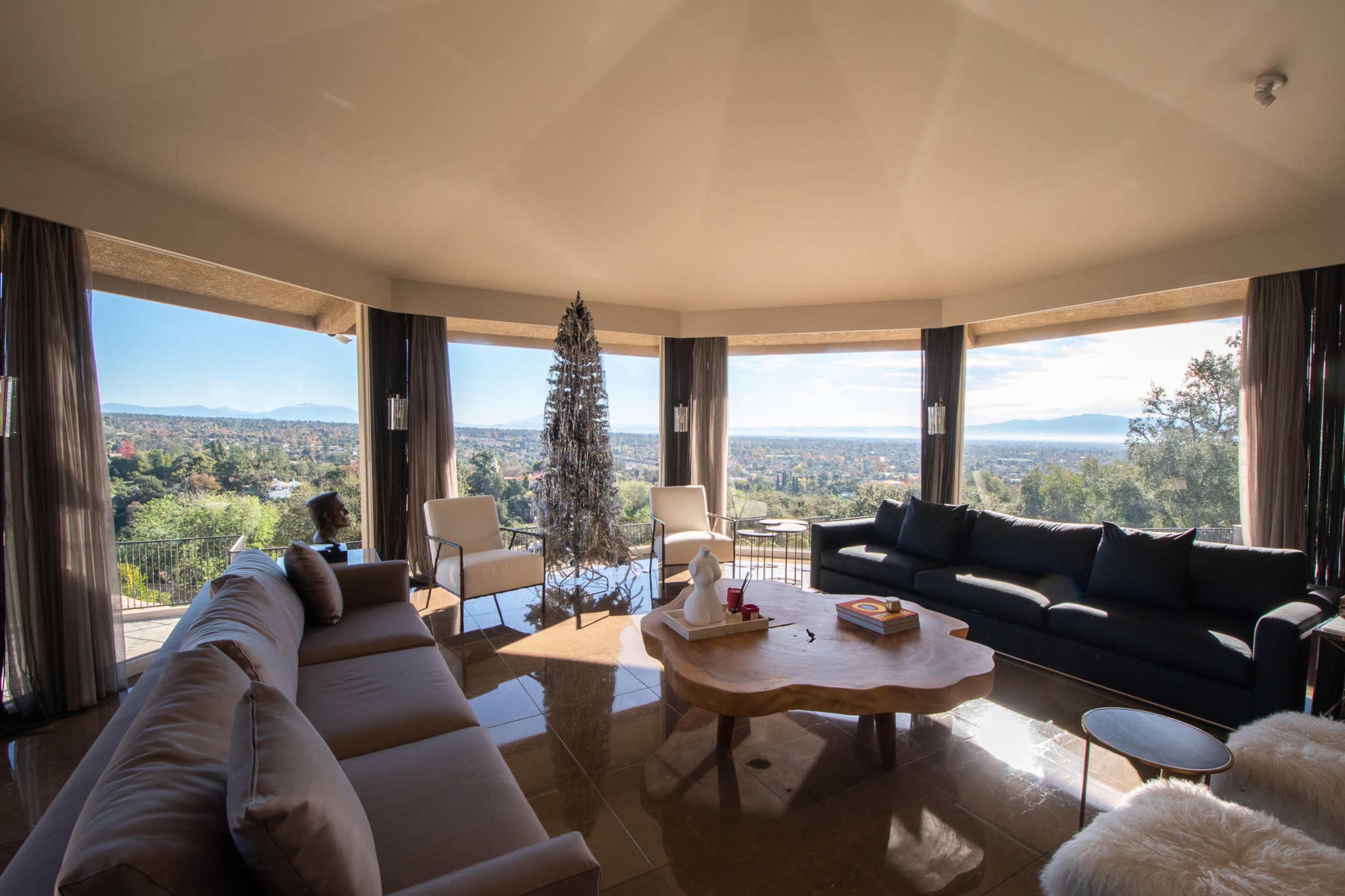 The image shows a modern living room with large windows offering a panoramic view of the landscape, featuring a coffee table made of wood and a tall decorative tree.
