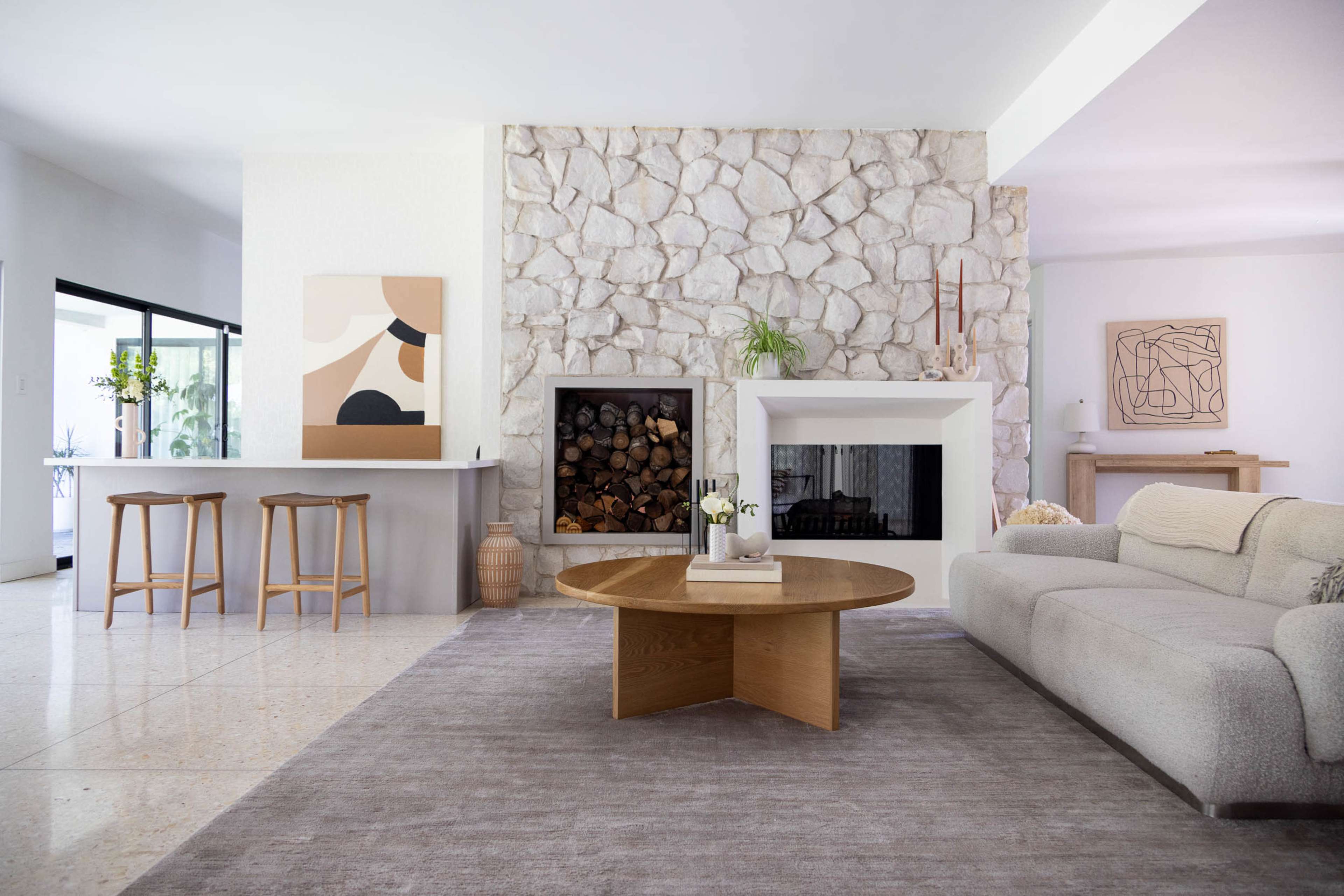 The image shows a modern living room featuring a stone fireplace, a round wooden coffee table, a light gray sofa, and a minimalist bar area with three stools.