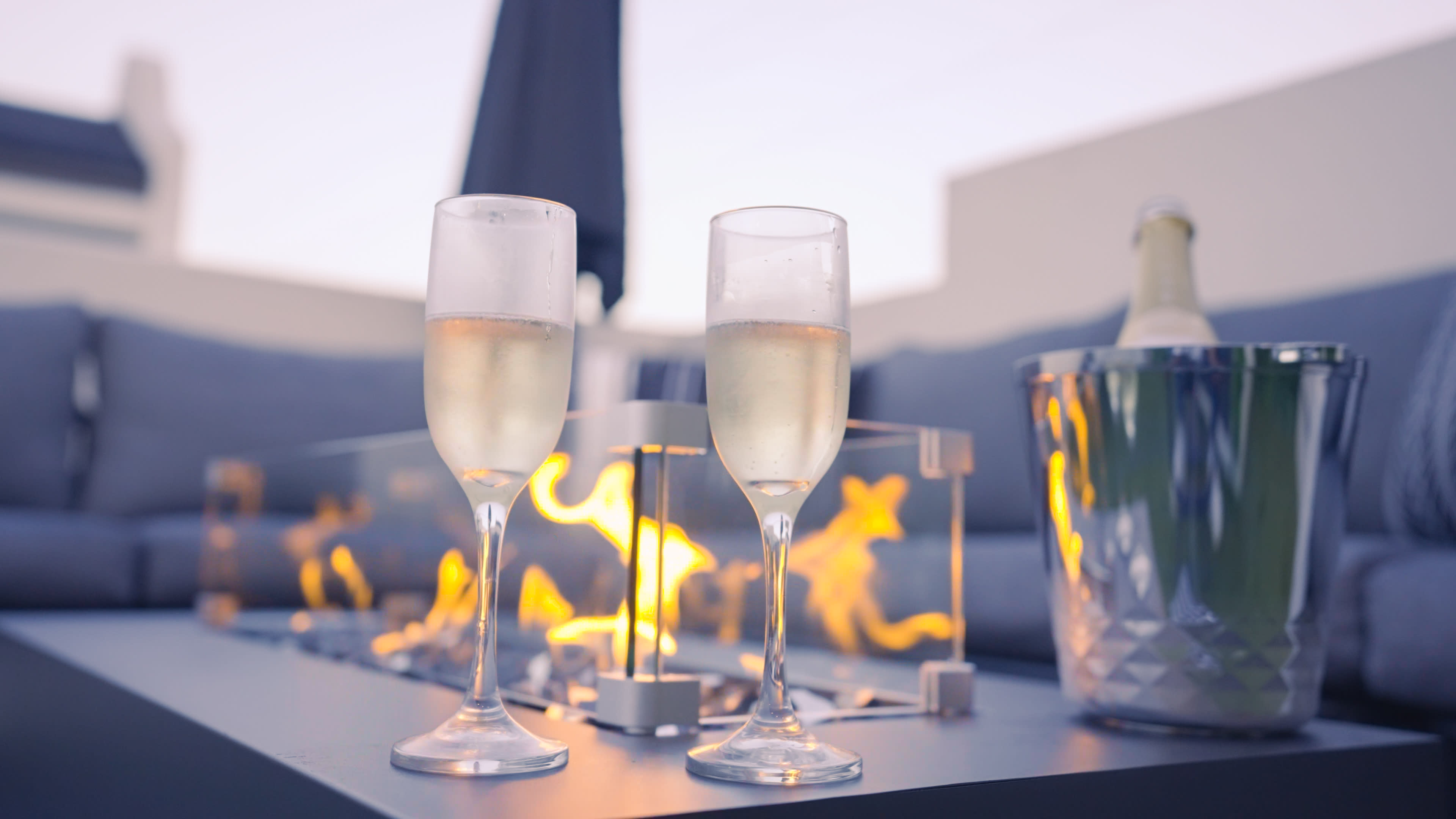 Two glasses of champagne sit beside a fire feature on a table, with a bottle of champagne in an ice bucket nearby, set on a patio.