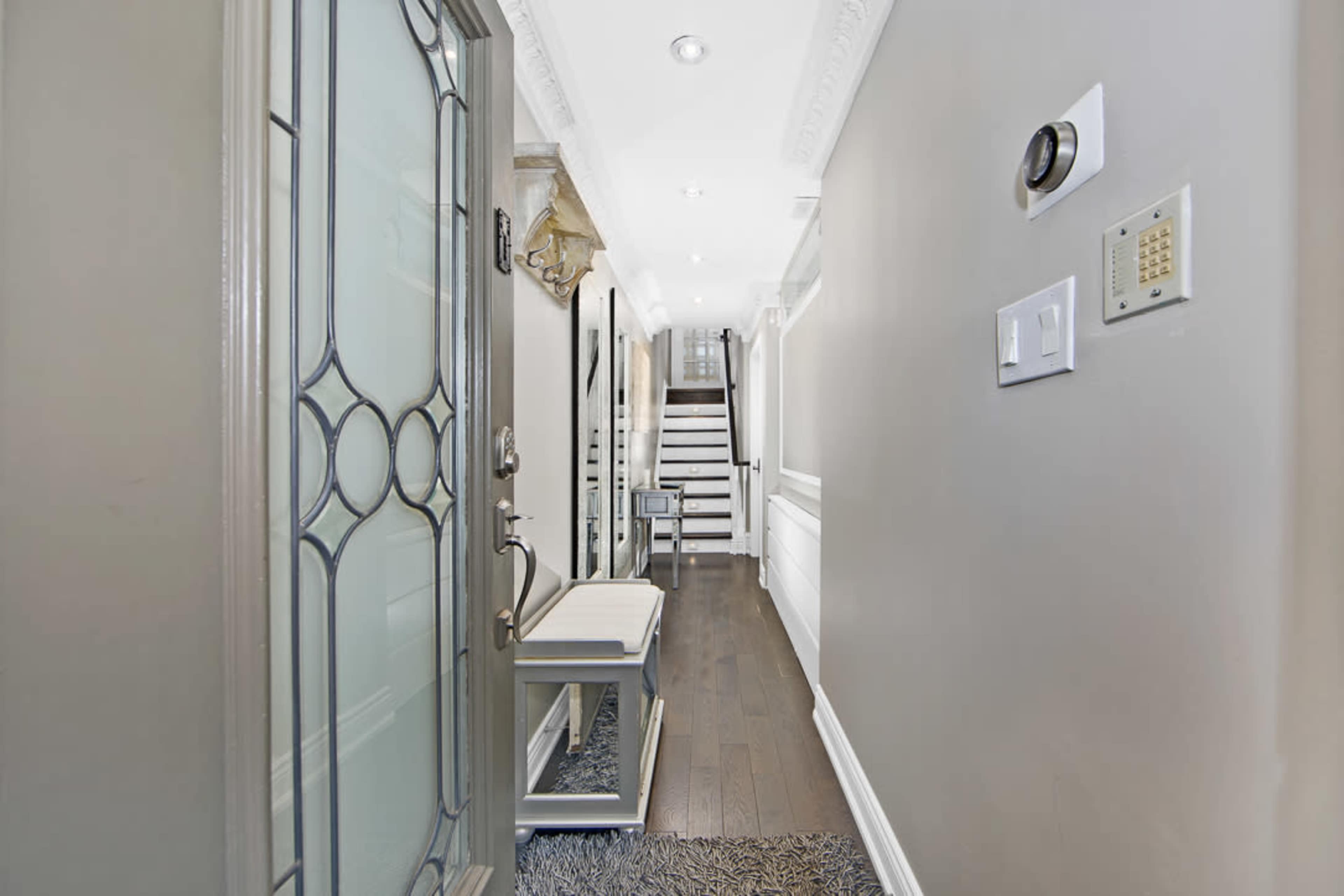 The image shows a well-lit hallway leading to a staircase, with a decorative front door and a bench along one side.