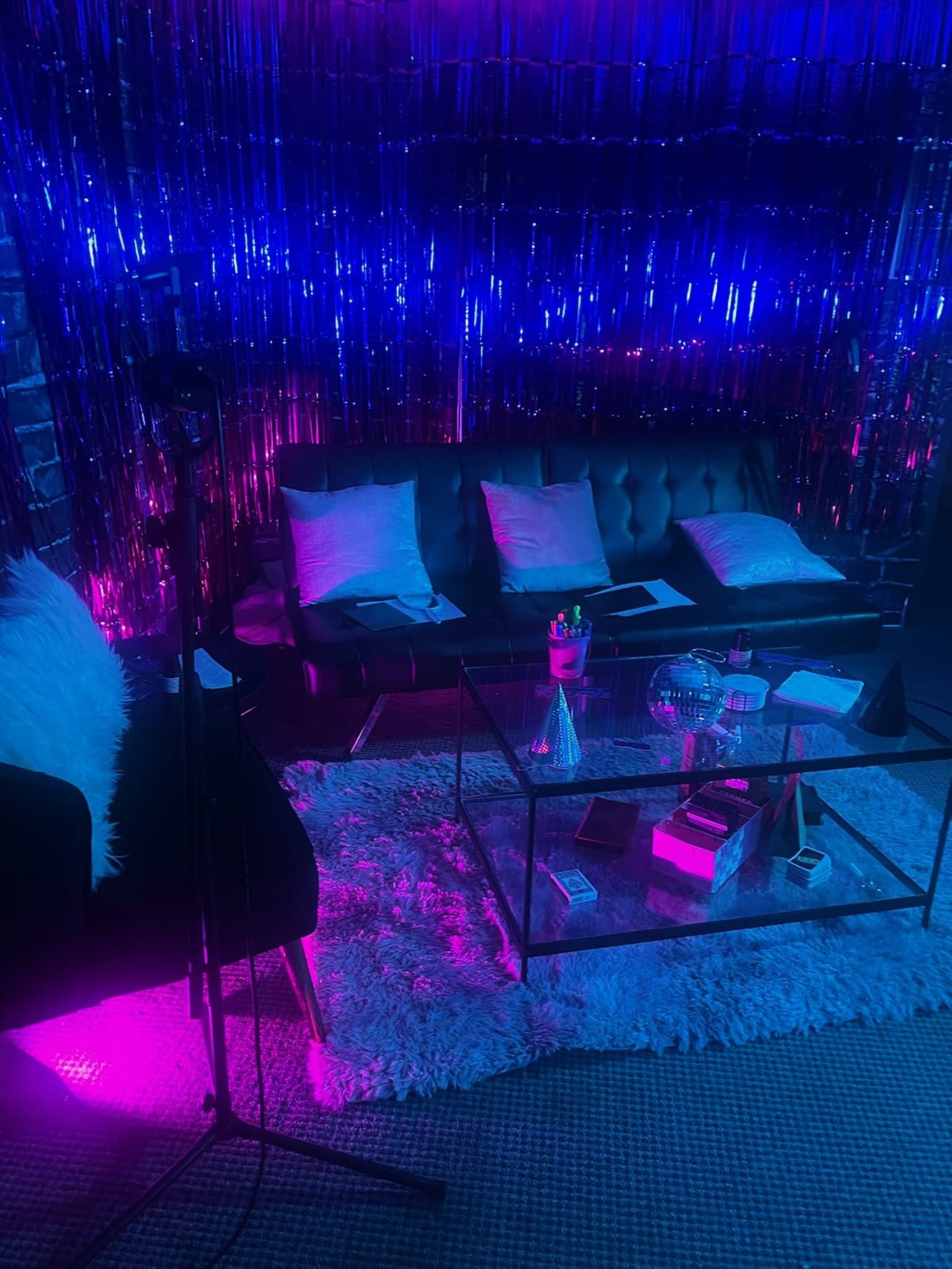 The scene features a modern lounge area with a black leather sofa, a glass coffee table, and colorful lighting effects created by shiny decorations in the background.