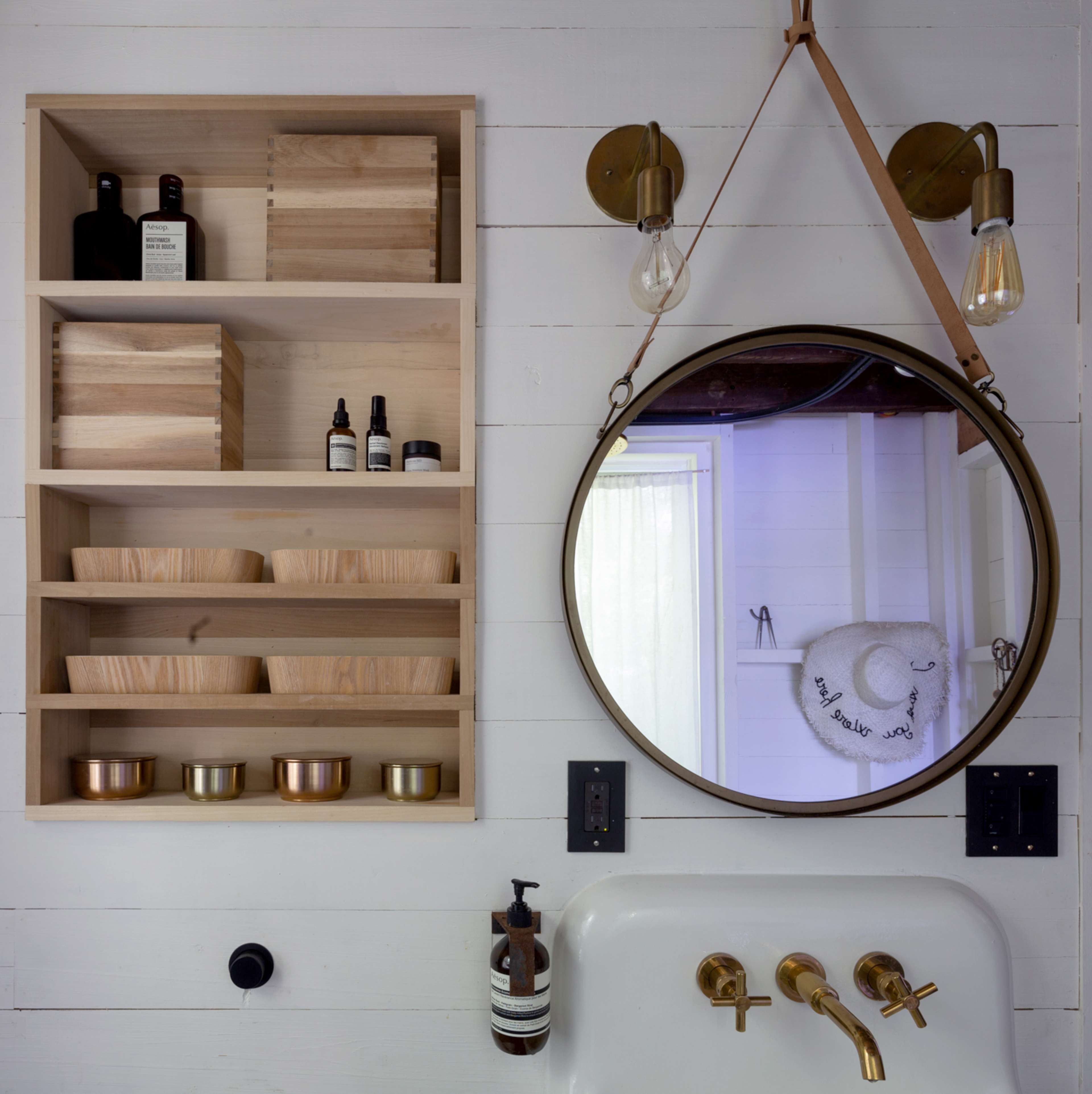 The image shows a neatly organized wall shelf with wooden boxes and containers above a sink featuring a round mirror and gold fixtures.