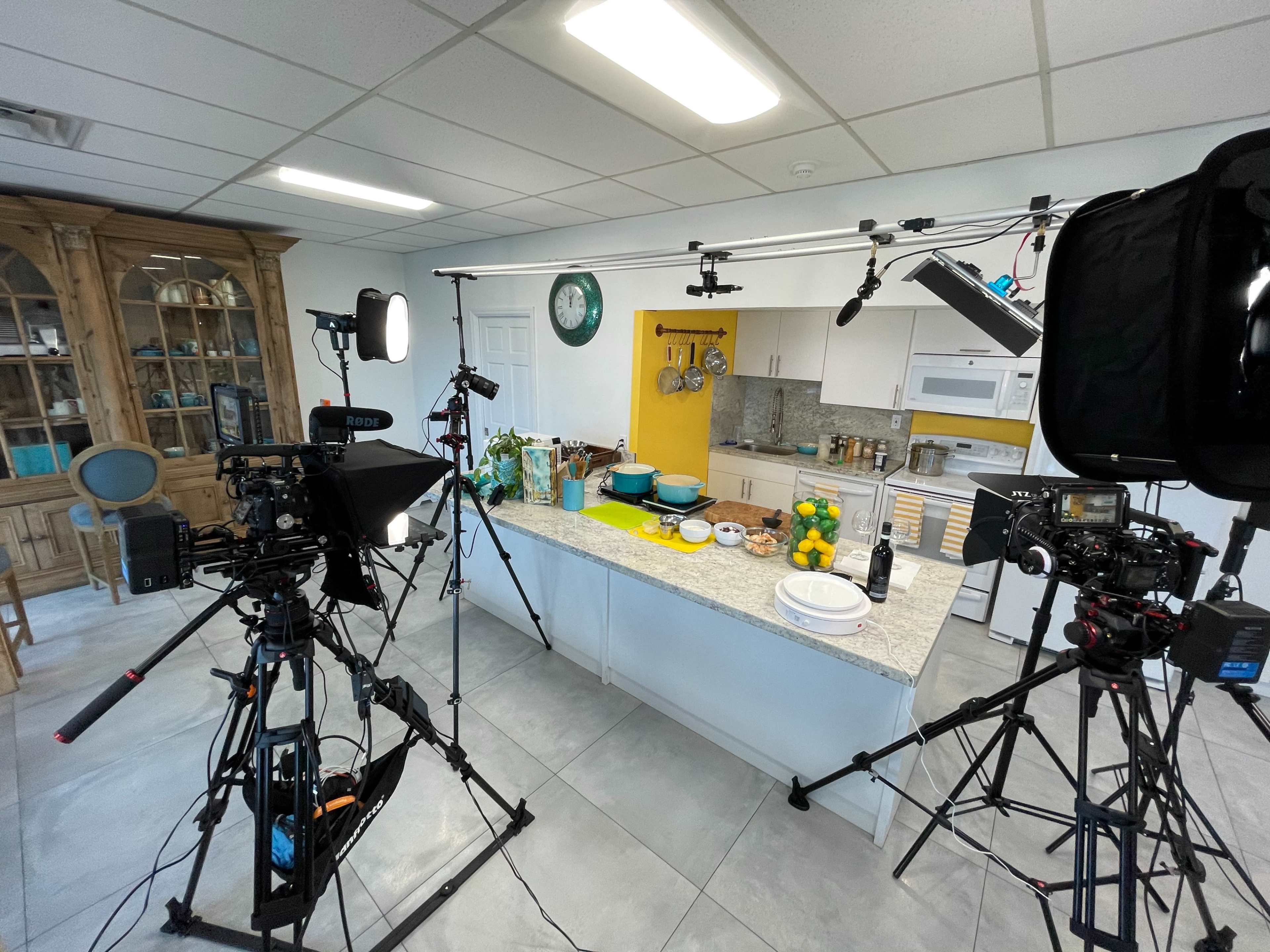 A kitchen setup for a filming session, featuring multiple cameras, lights, and a table with various food items.