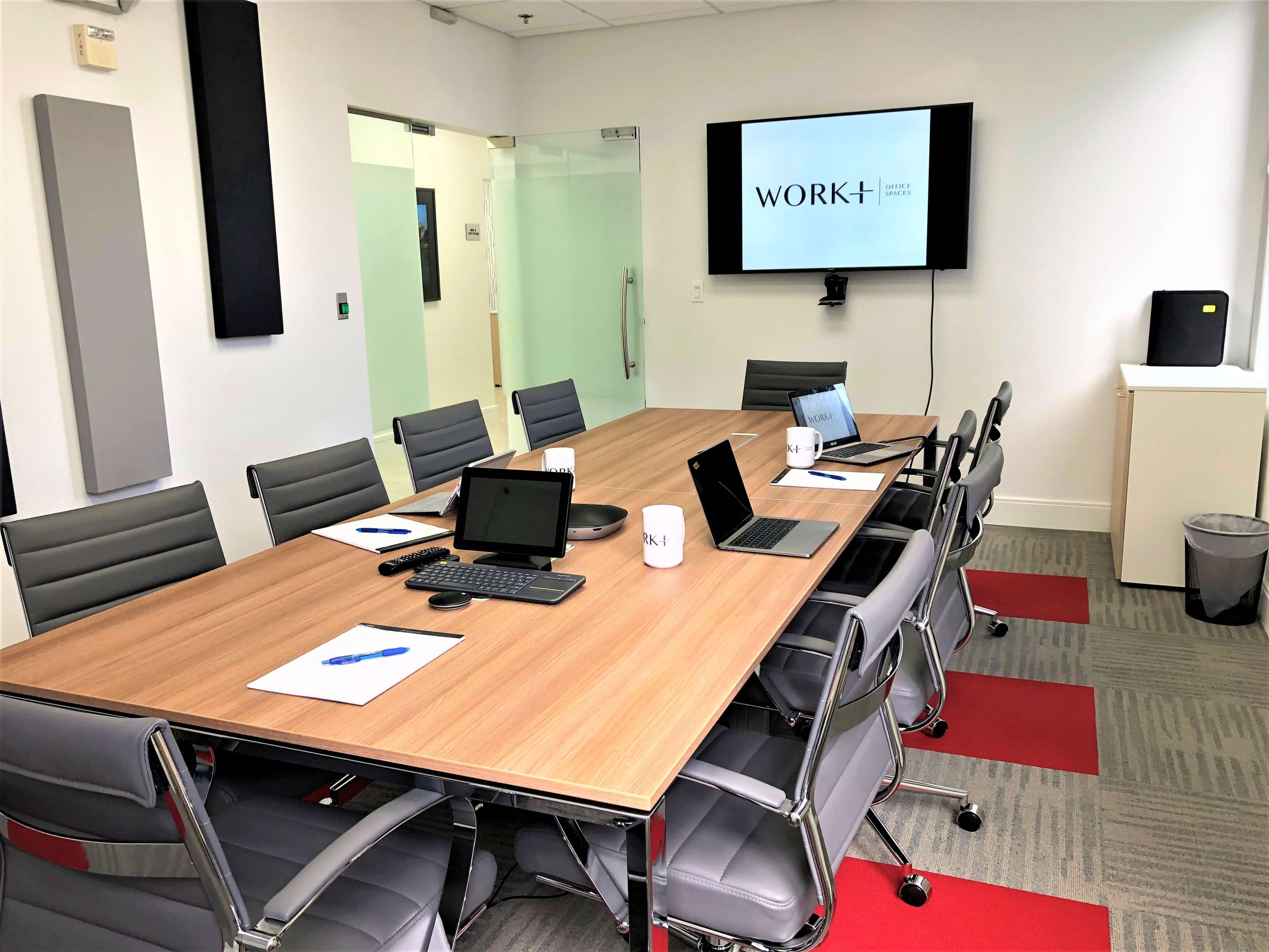 A modern conference room is set up with a long wooden table, multiple laptops, notepads, and a large screen displaying the word "WORK+" on the wall.
