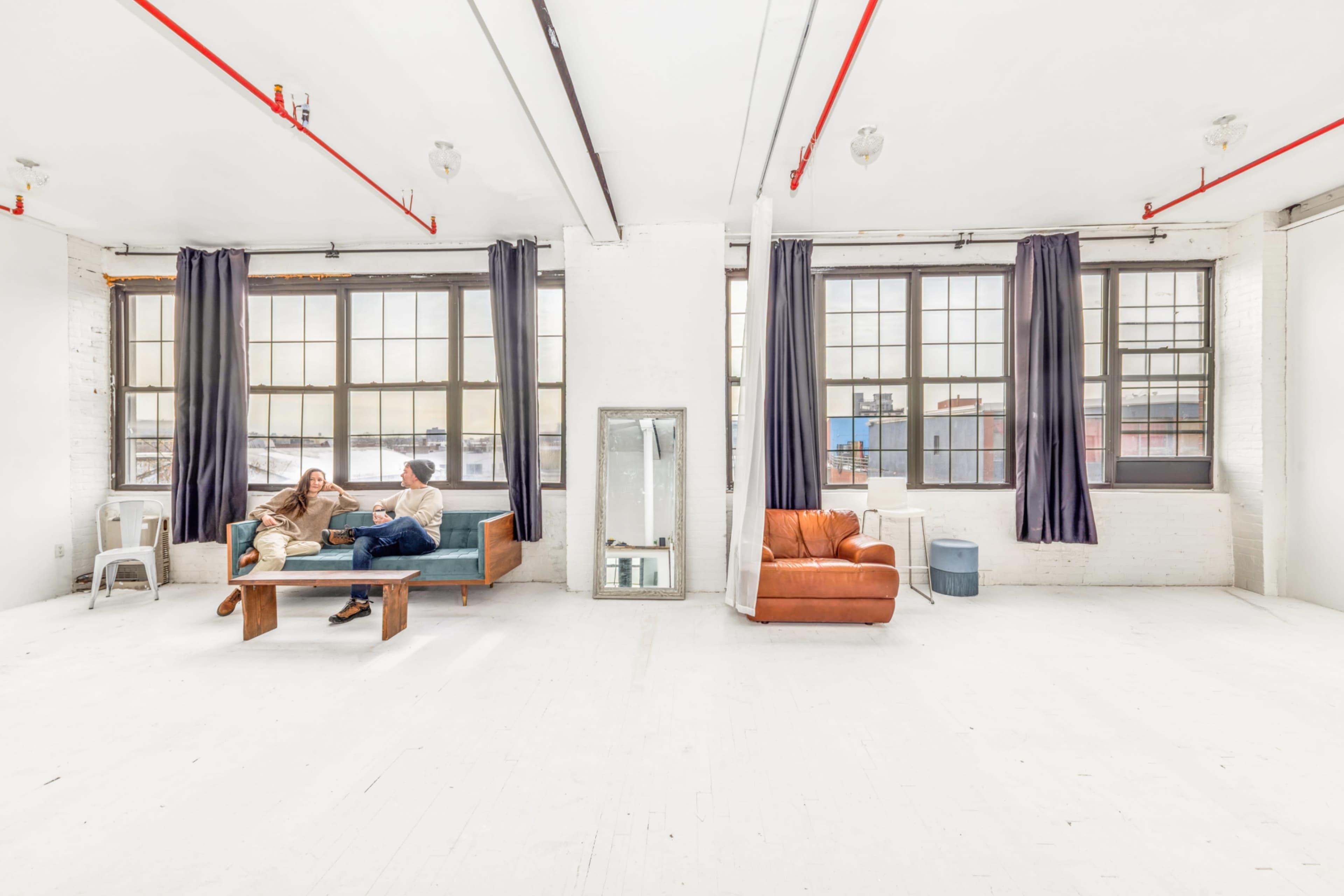 A bright, spacious room features large windows, two people sitting on a couch, and minimal furniture.