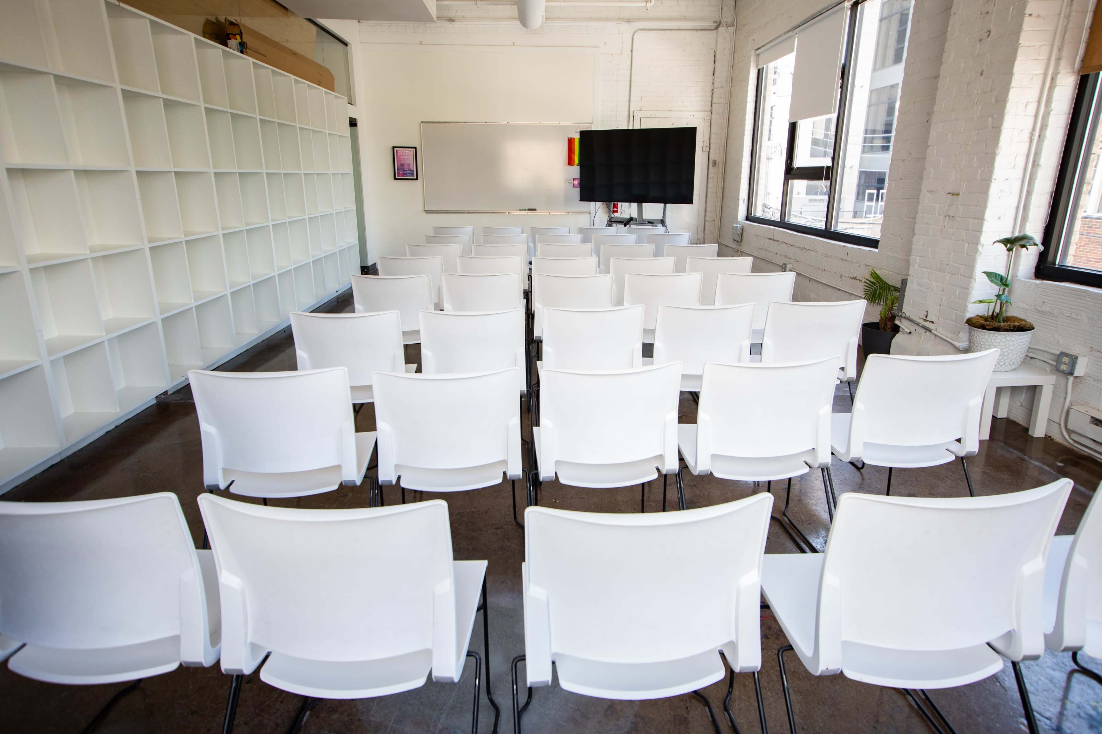 The image shows a classroom with rows of empty white chairs facing a blank whiteboard and a large screen in a well-lit room.