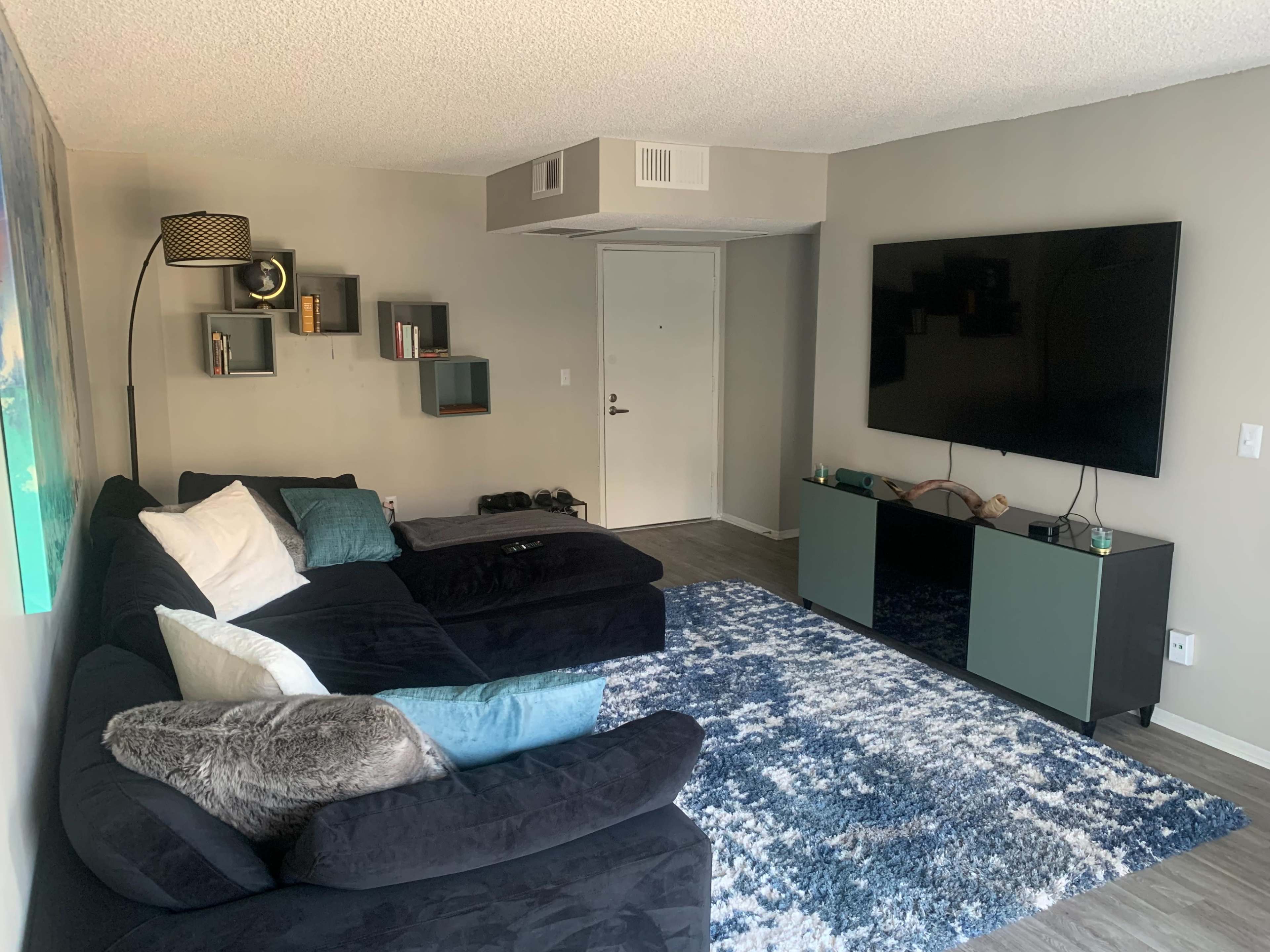 A modern living room featuring a large black sectional sofa, a flat-screen TV mounted on the wall, and a light blue area rug on a wooden floor.