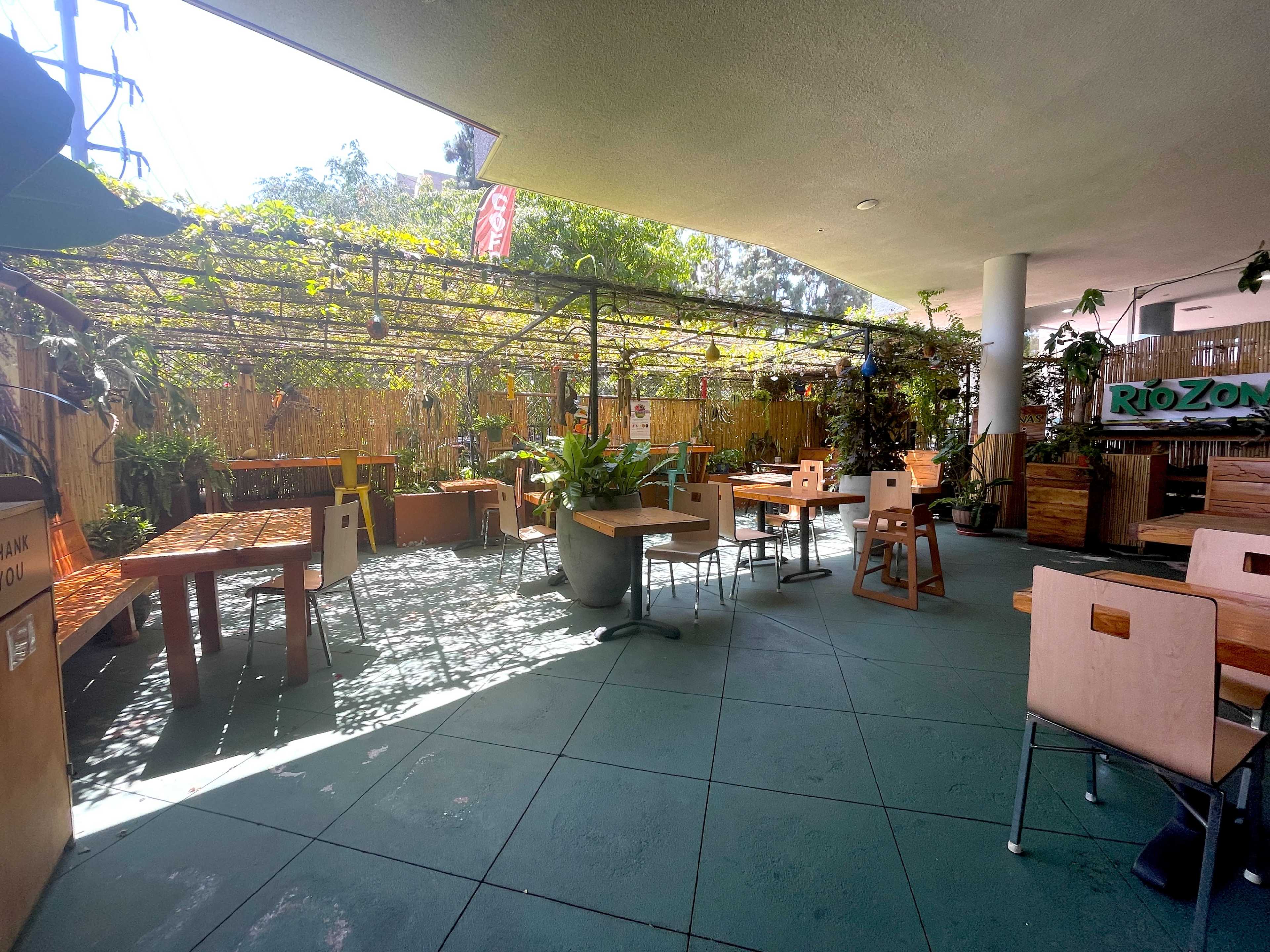 The image shows a partially shaded outdoor dining area with wooden tables and chairs surrounded by greenery and plants.