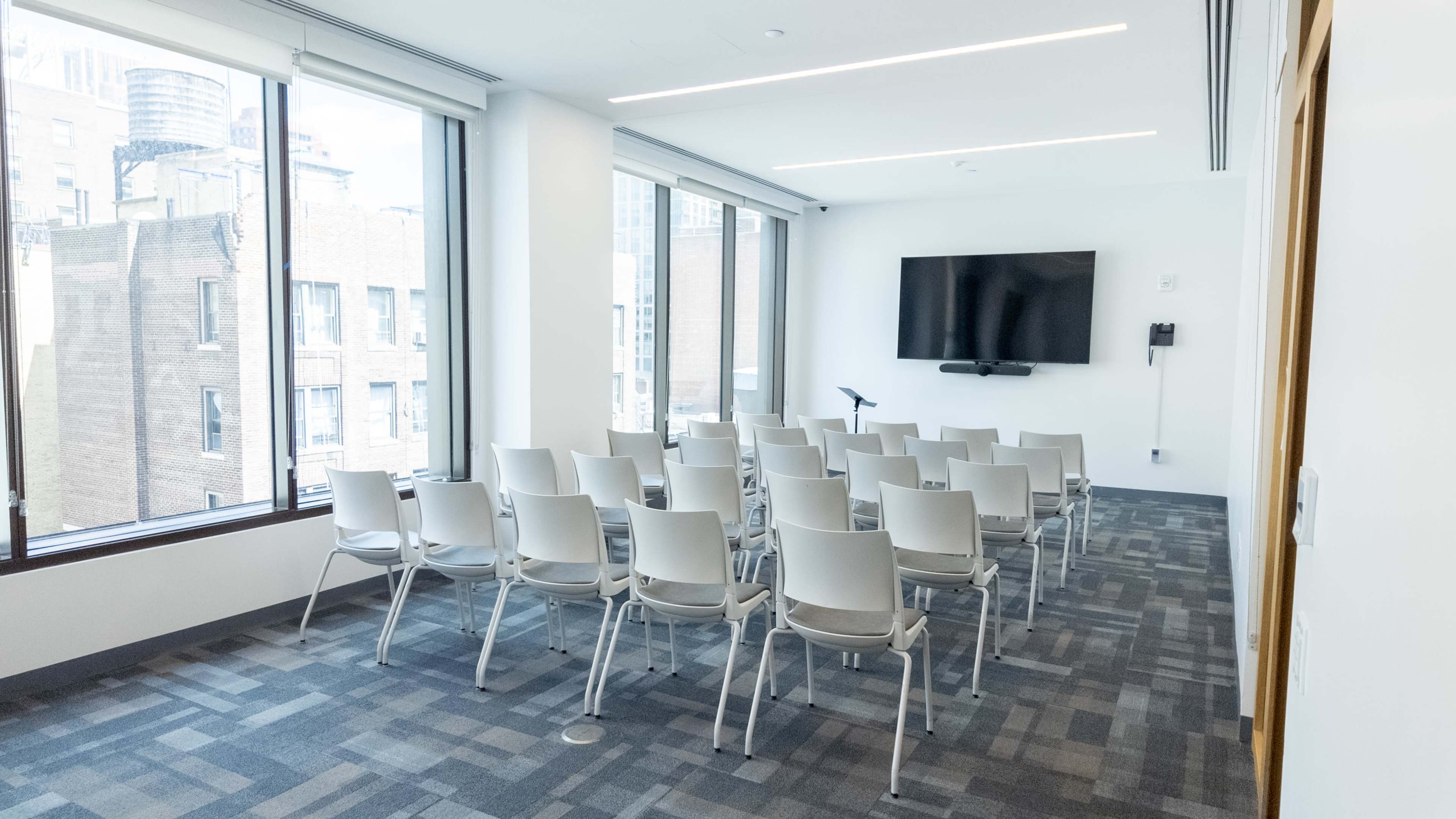 The image shows a modern conference room with rows of white chairs facing a large wall-mounted screen and tall windows overlooking a cityscape.