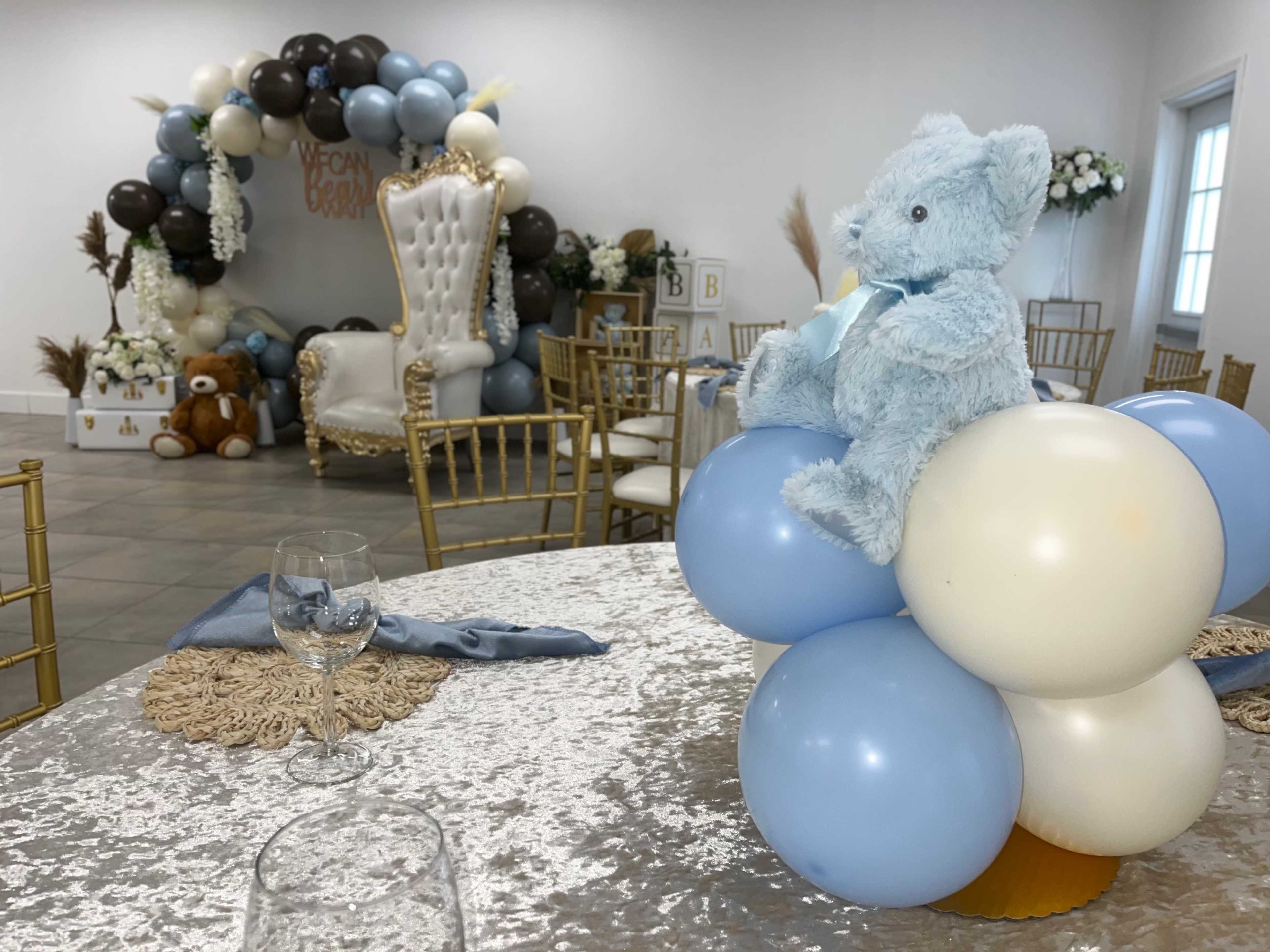 A decorated event space features a large teddy bear centerpiece atop balloons, with a backdrop of balloons and a throne chair in the background.