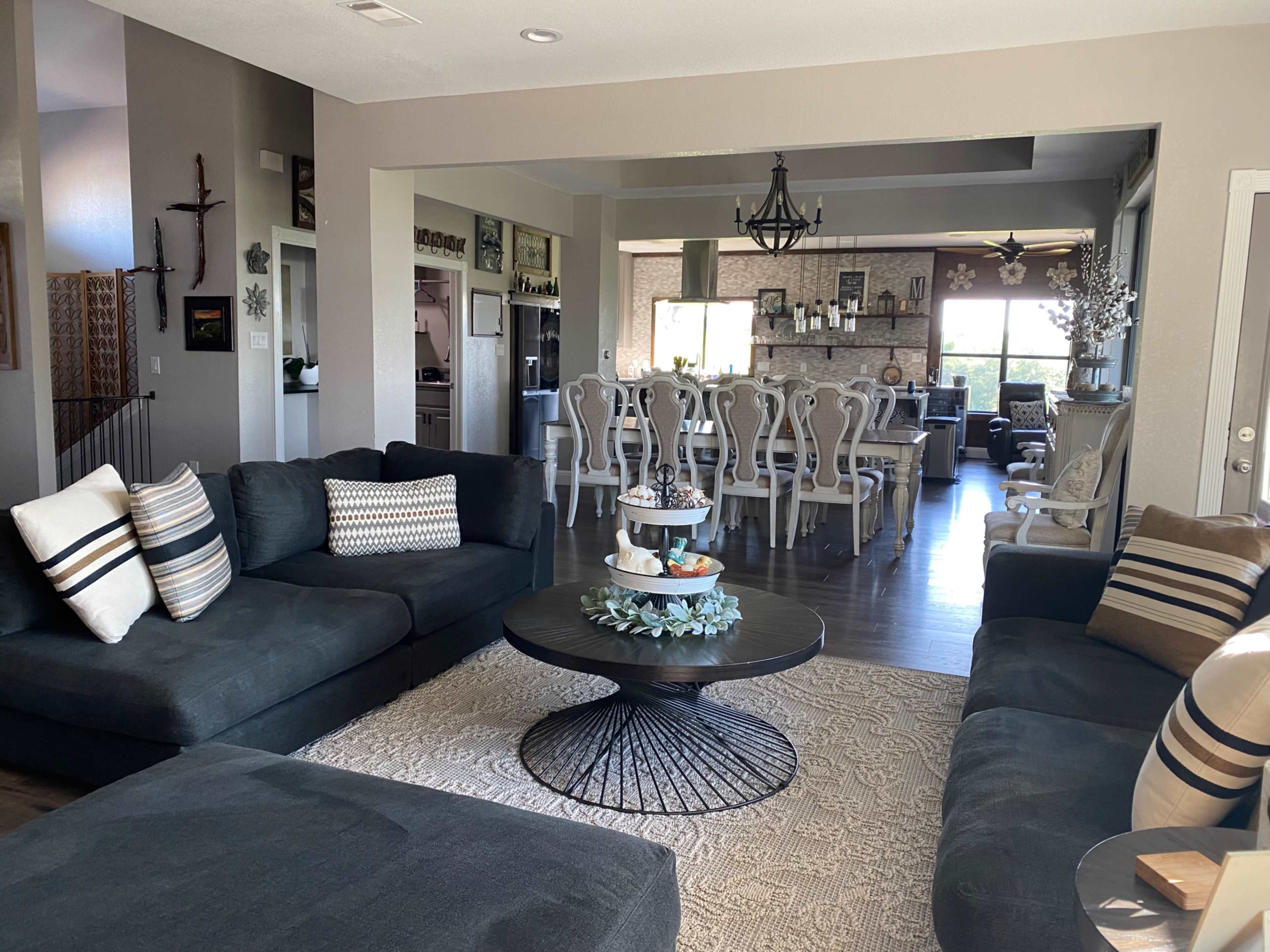A spacious living room with dark sectional sofas, a round coffee table, and a decorative area rug, leading into a dining area with white chairs and a large table.