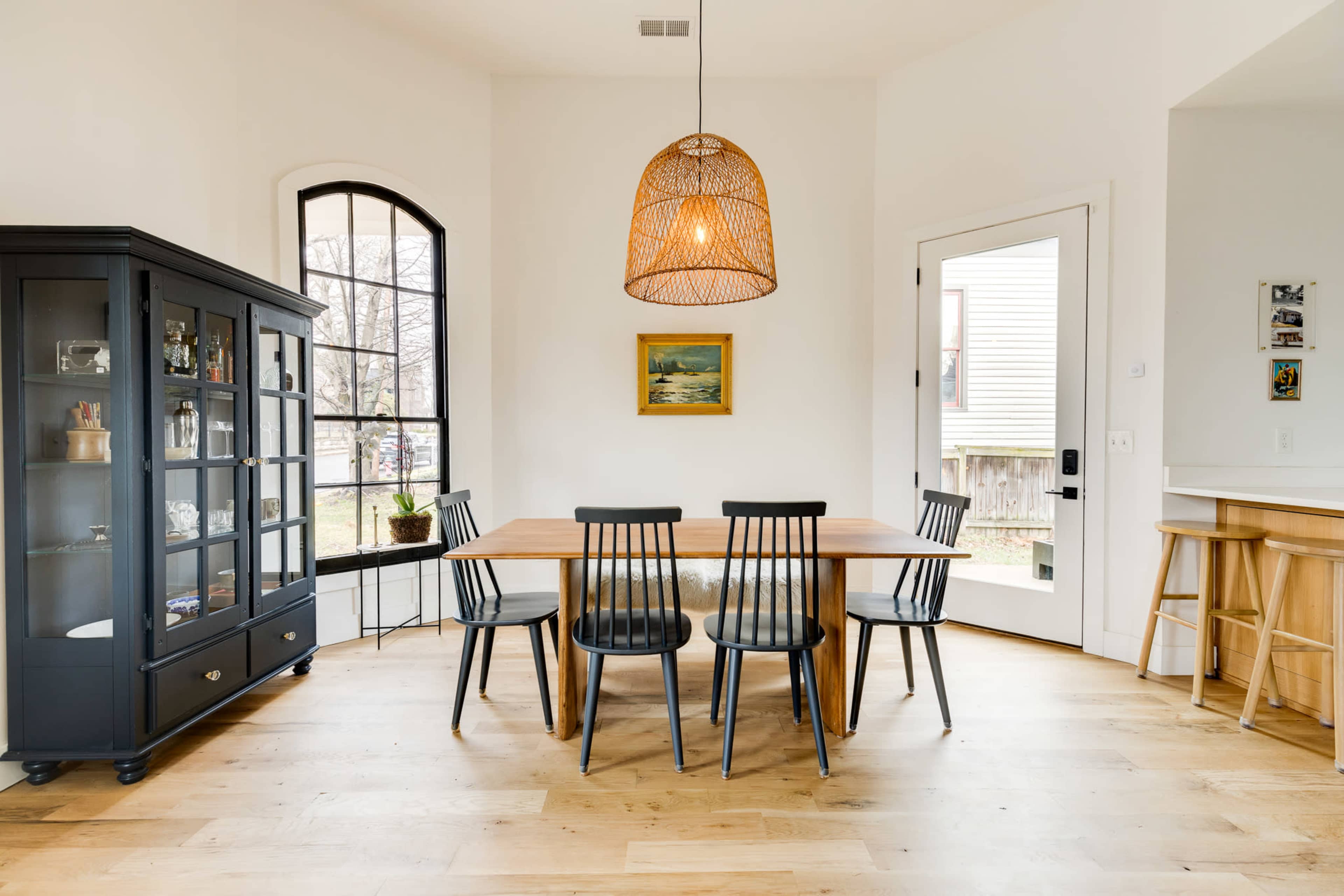 A simple dining area features a wooden table surrounded by black chairs, with a window and a door leading outside, alongside a glass-fronted cabinet.