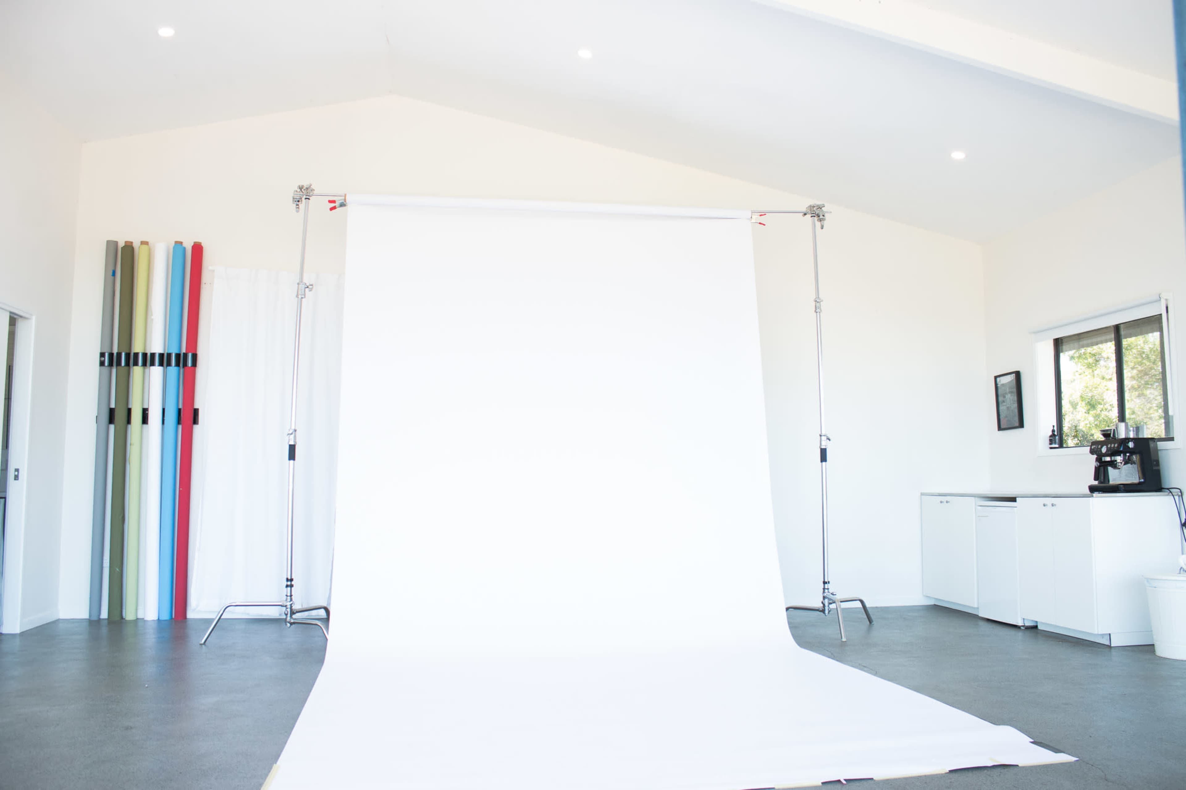 The image shows a photography studio with a large white backdrop set up against a light-colored wall and various colored panels leaning on the side.