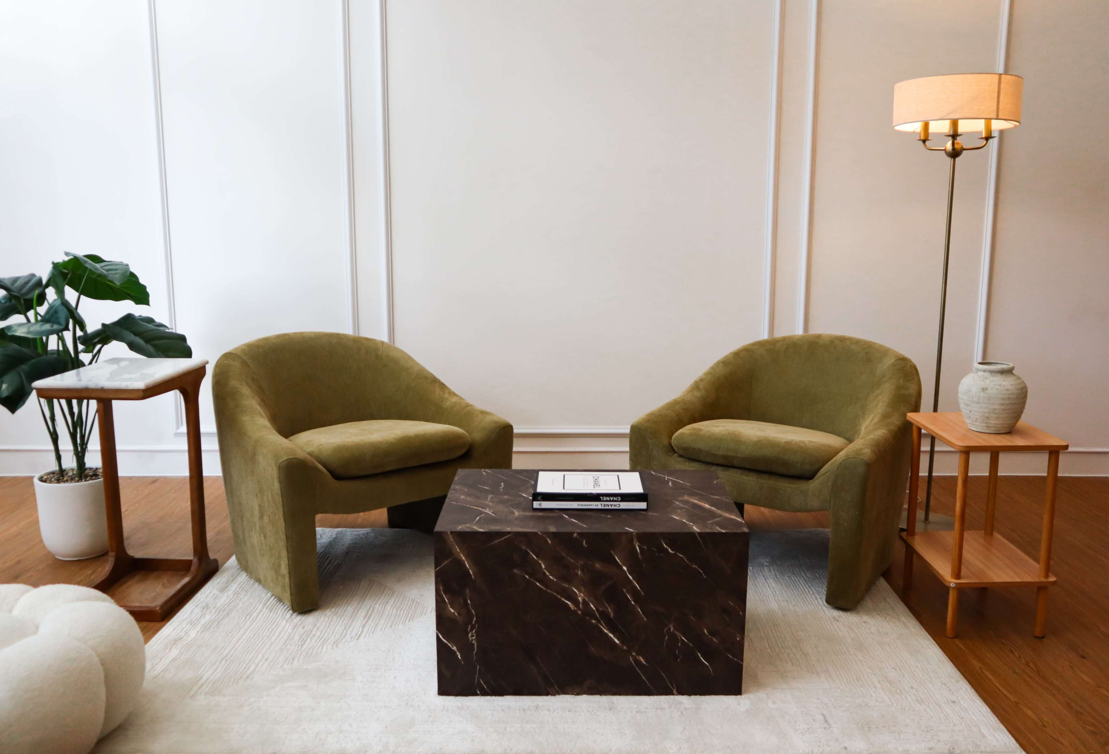The image shows a minimalist living room with two green chairs facing a dark marble coffee table, accompanied by a floor lamp and a potted plant on a small side table.