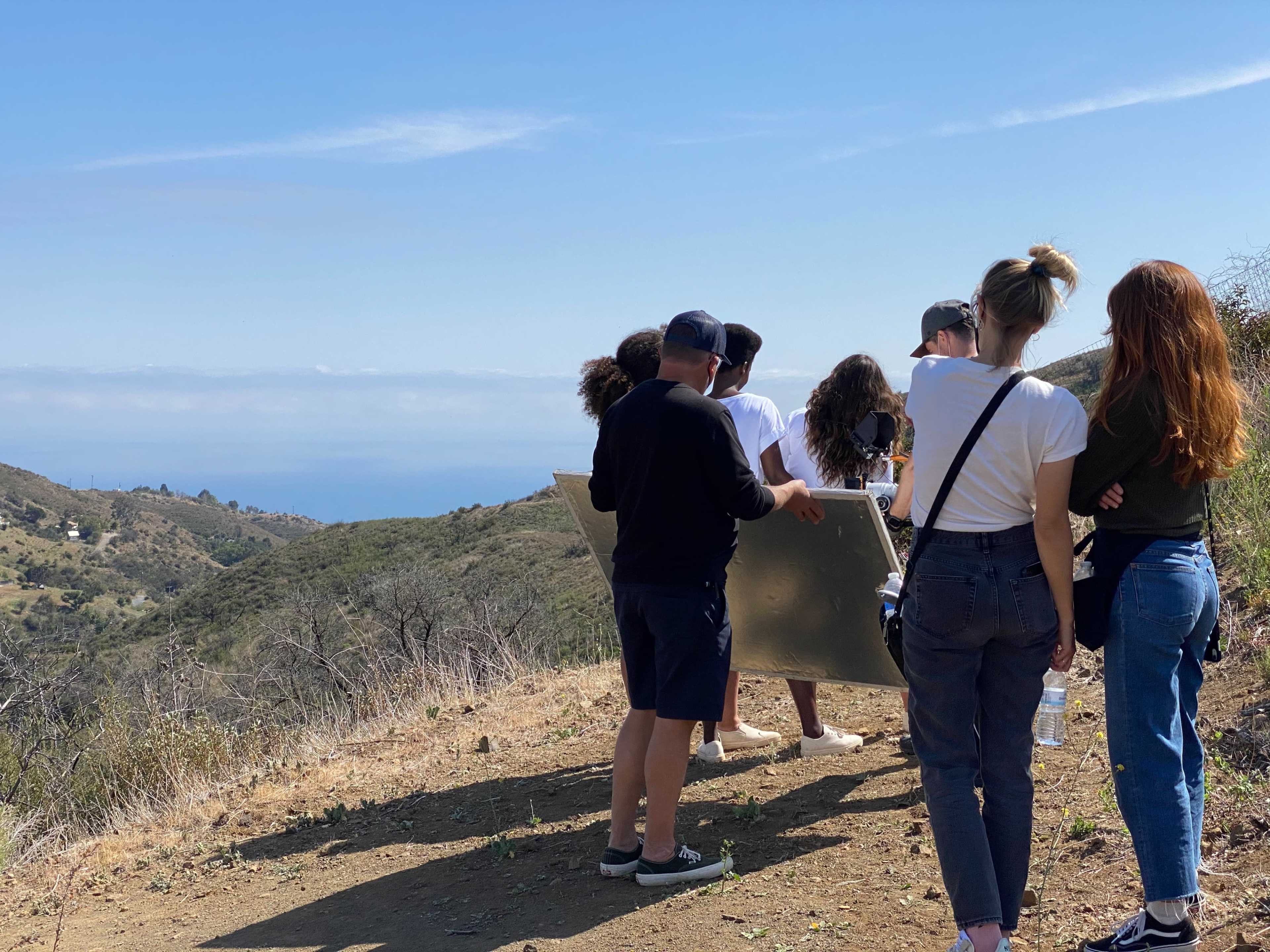 A group of people stands on a hillside overlooking a body of water in the distance, with mountains and greenery surrounding them.