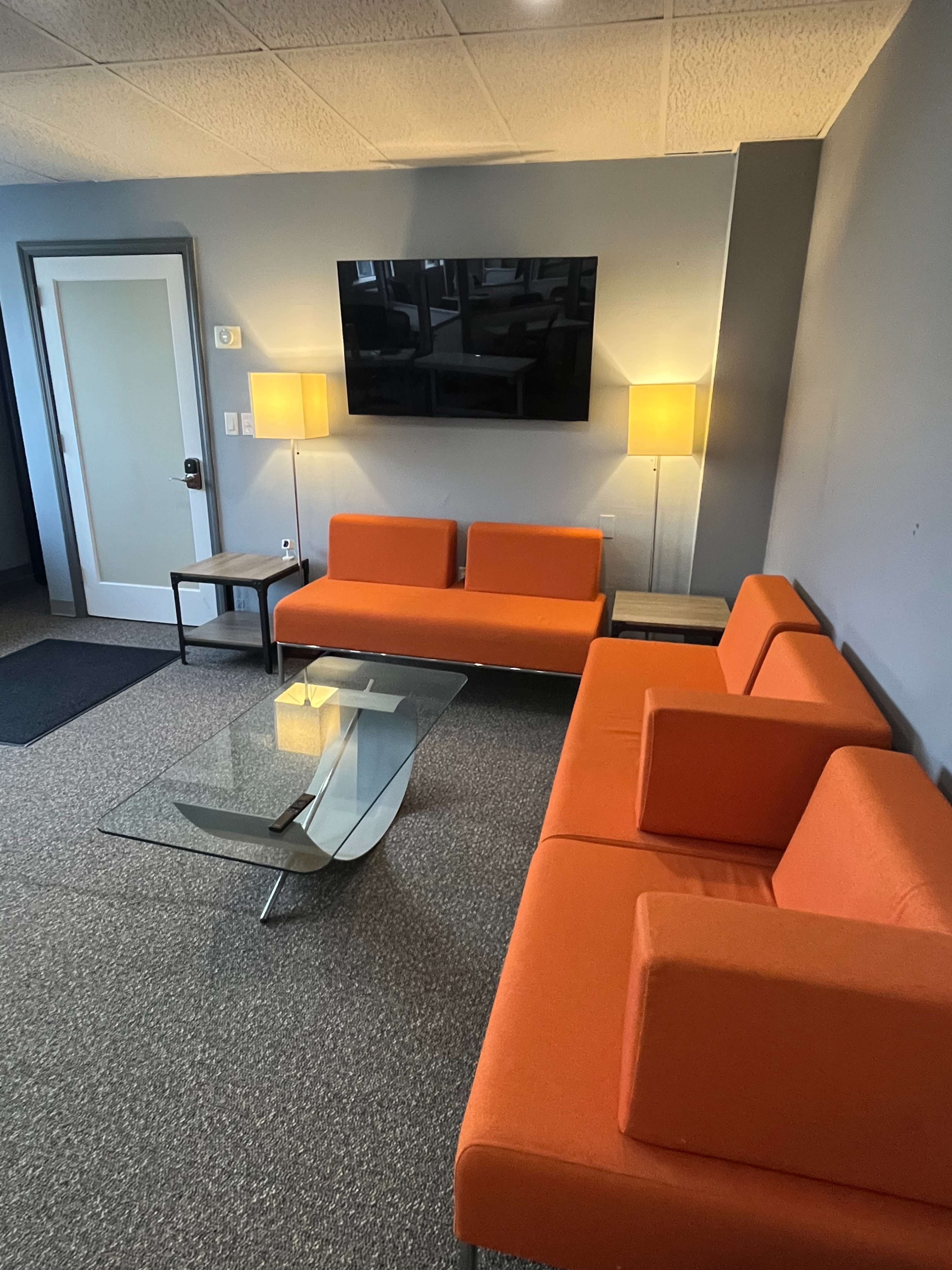 The image shows a modern lounge area with orange couches, a glass coffee table, two lamps, and a wall-mounted television.