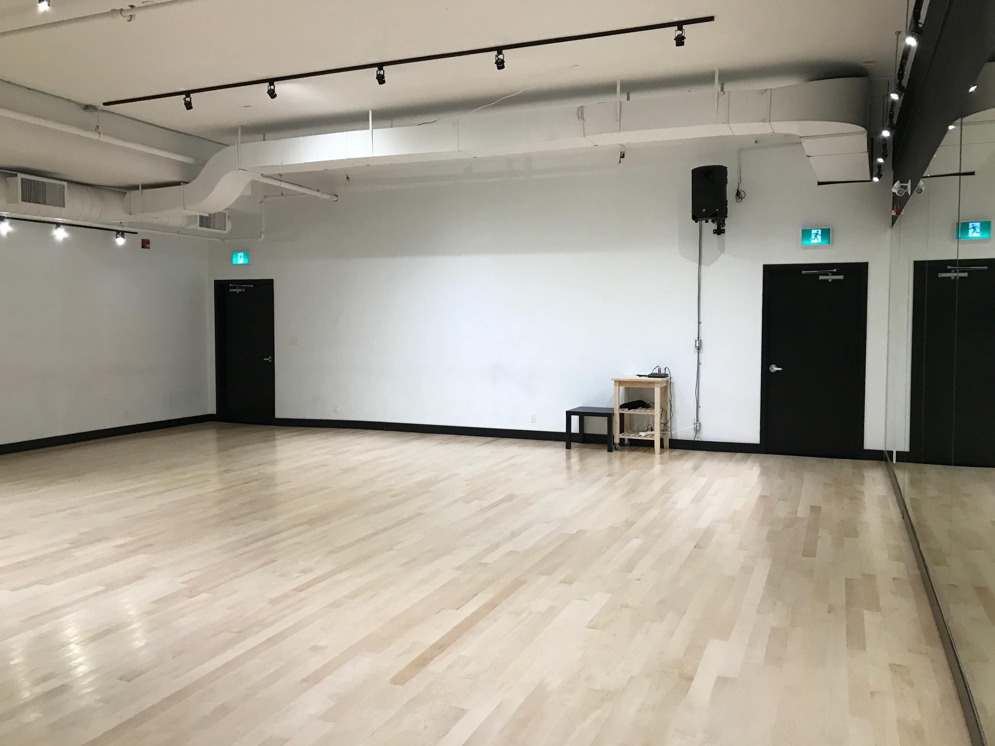 The image shows a spacious, empty room with a polished wooden floor, mirrored walls, and black doors on either side.