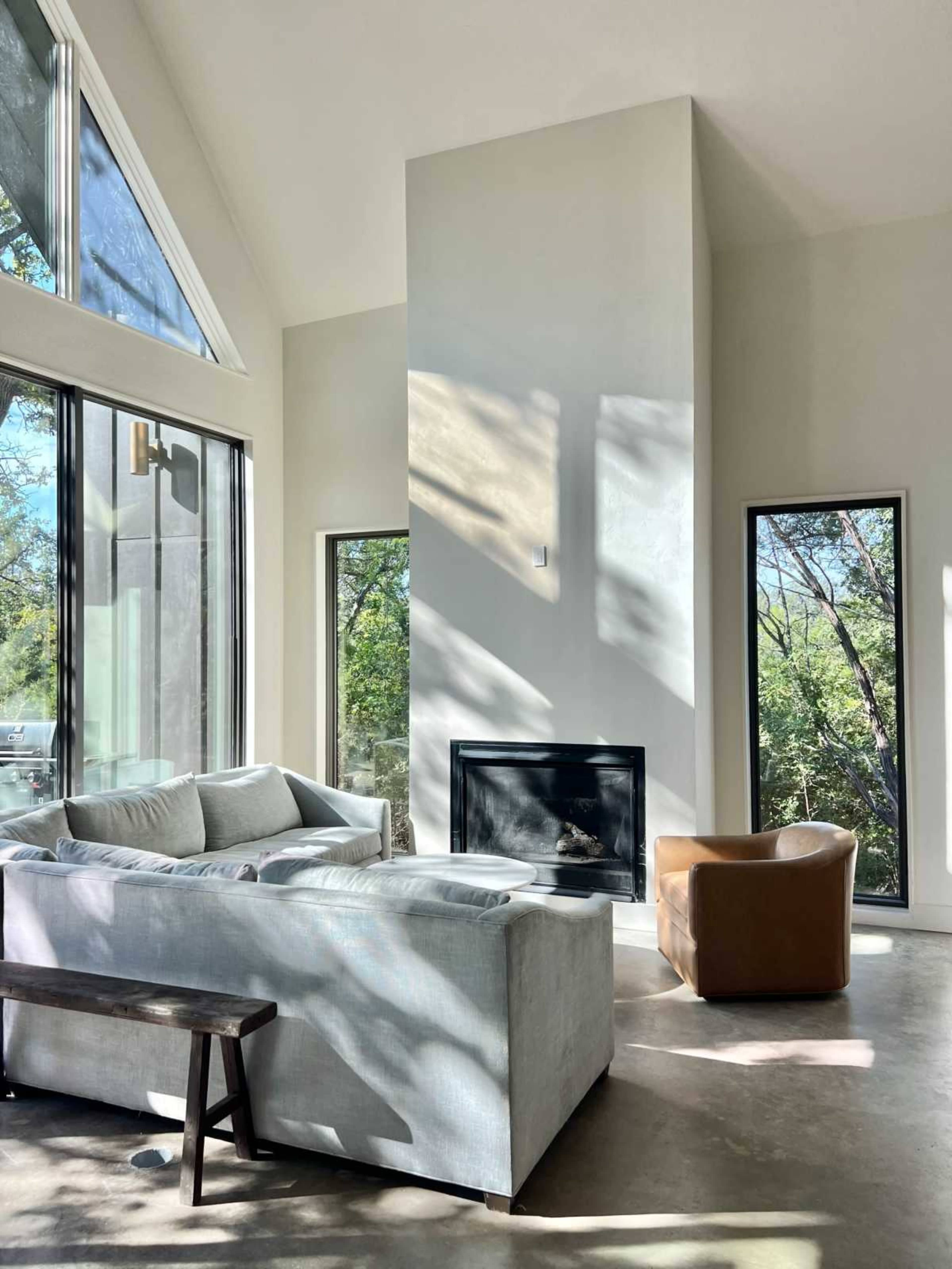 The image shows a modern living room with a light-colored sofa, a brown chair, and large windows allowing natural light to illuminate the space.