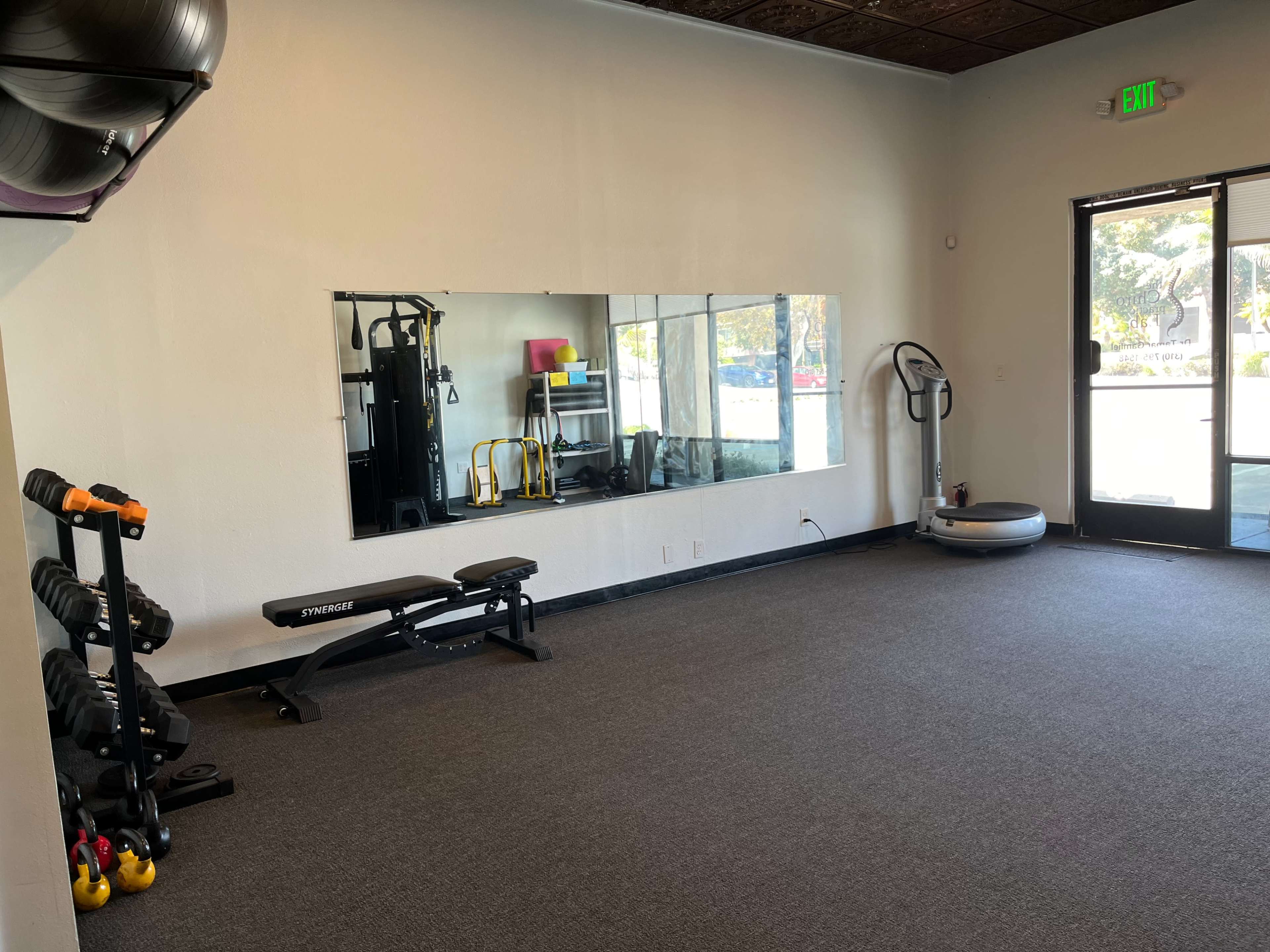 The image shows an empty gym with exercise equipment, including dumbbells, a weight bench, and a mirror along one wall.