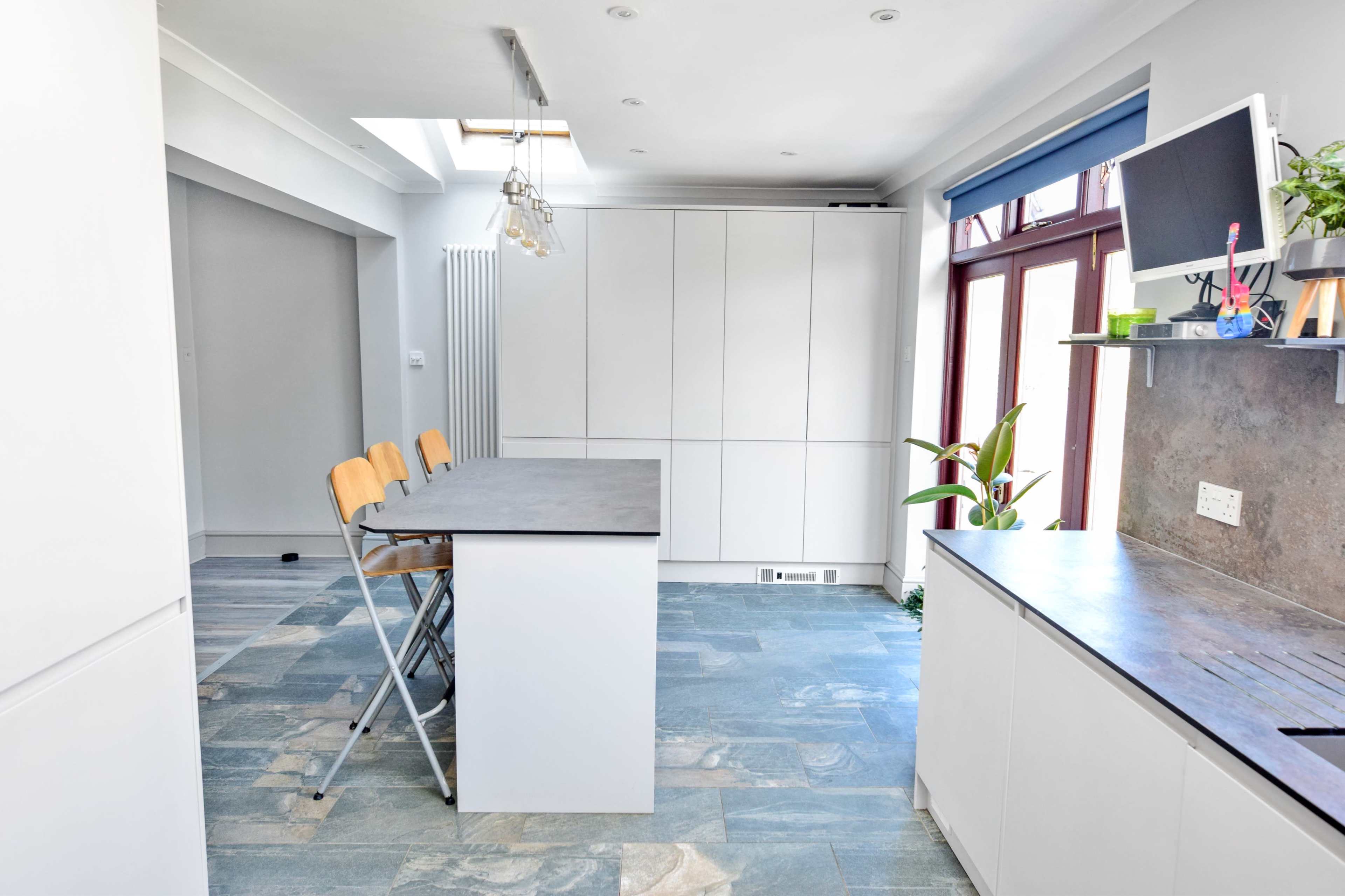A modern kitchen features a central island with bar stools, white cabinetry, and large windows allowing natural light to enter.