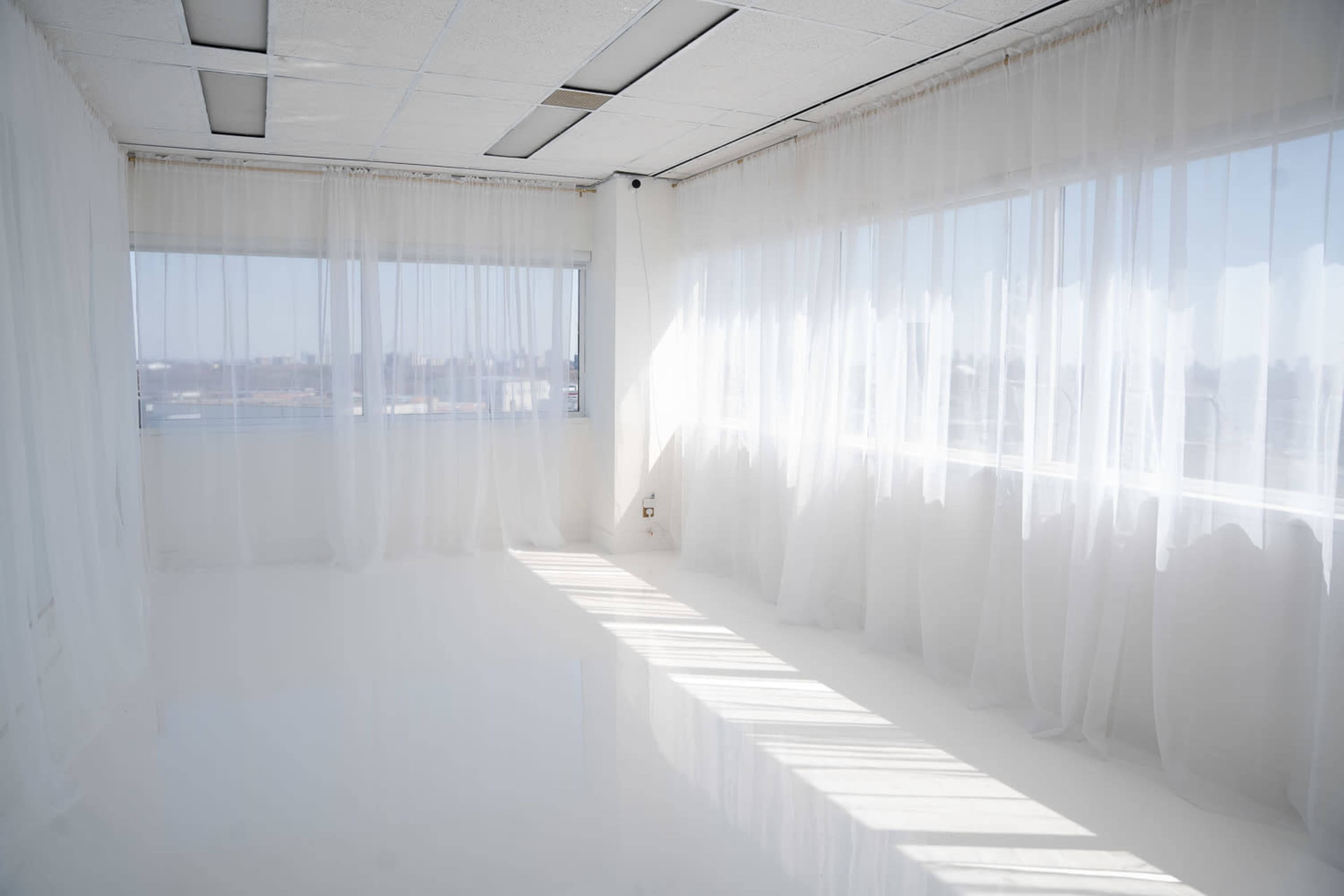 The image shows a brightly lit room with white walls, a glossy floor, and sheer white curtains framing large windows.