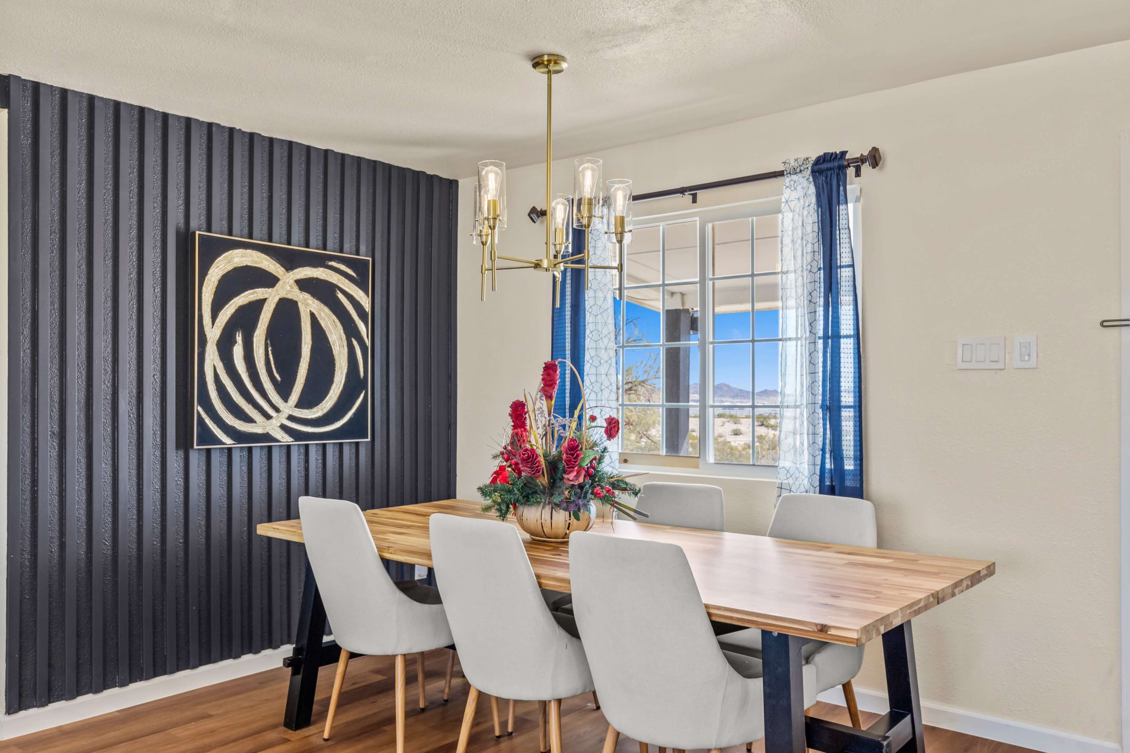 A dining area features a wooden table surrounded by light-colored chairs, with a decorative floral centerpiece and a window offering a view of the outdoors.
