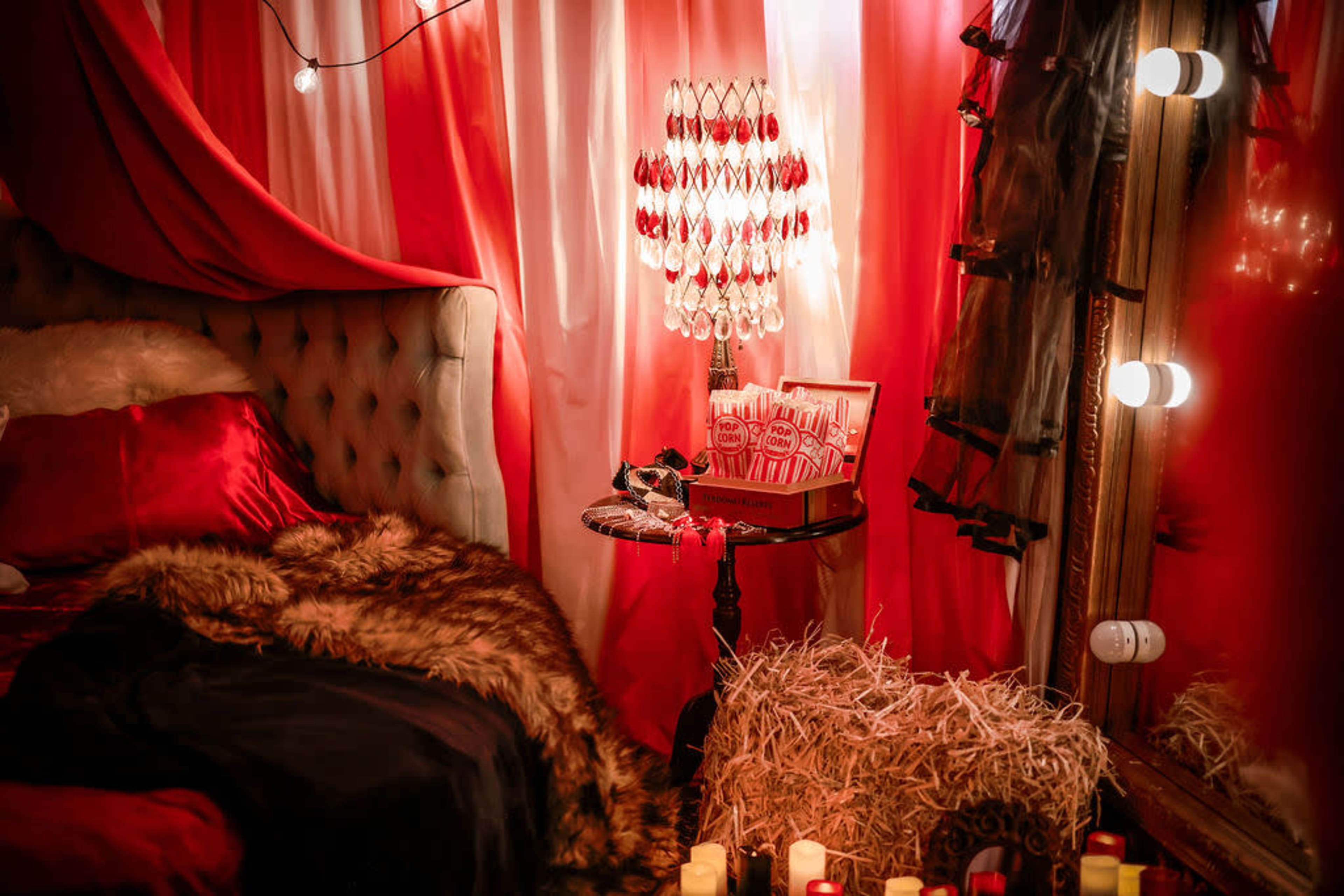 The image shows a cozy bedroom corner adorned with red and white drapes, a vintage-style lamp, and decorative elements like a fur throw and a small table with candy.