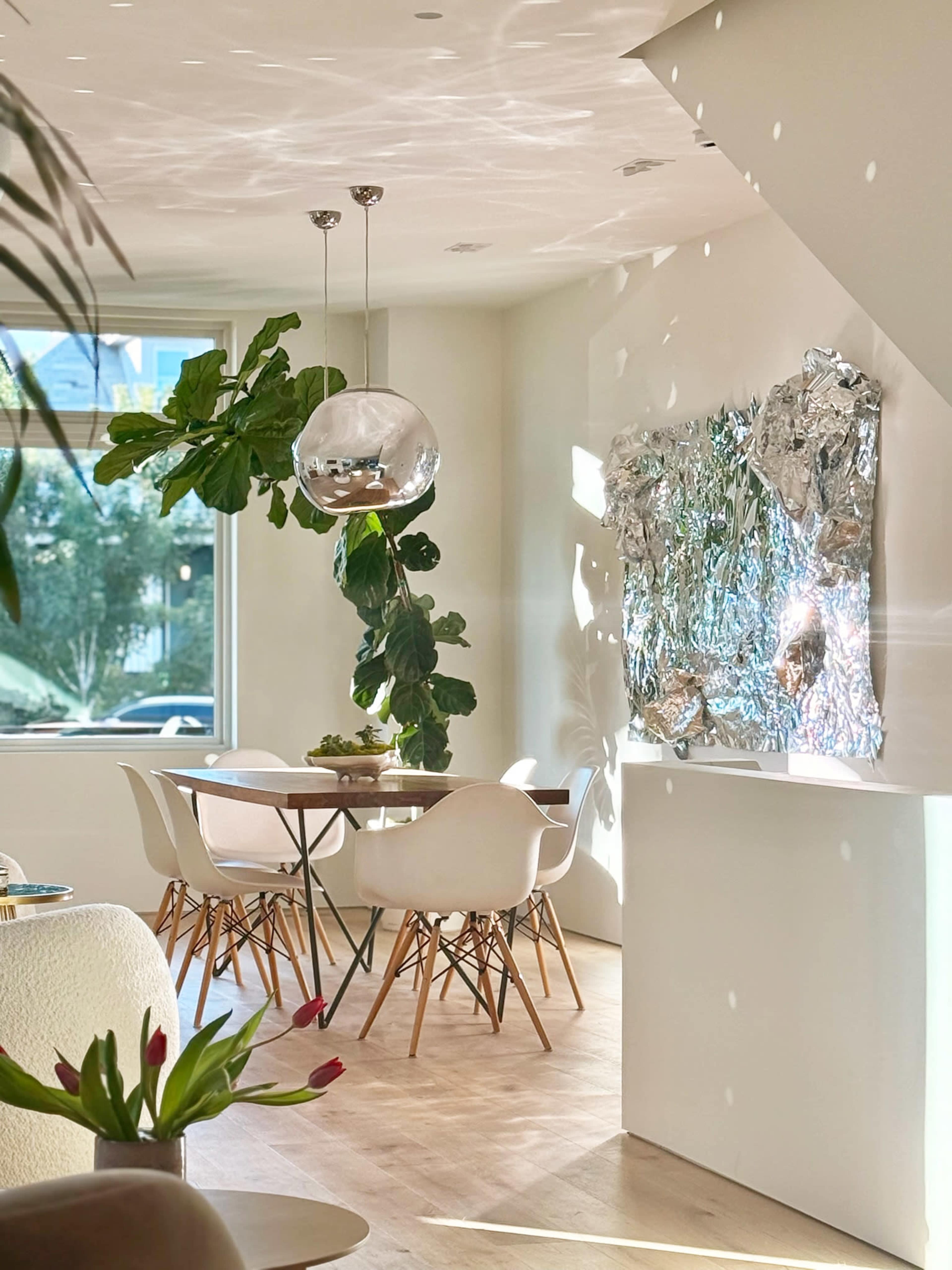 A modern dining area features a round table surrounded by white chairs, illuminated by natural light filtering through windows, with a decorative metallic wall piece and a hanging plant.