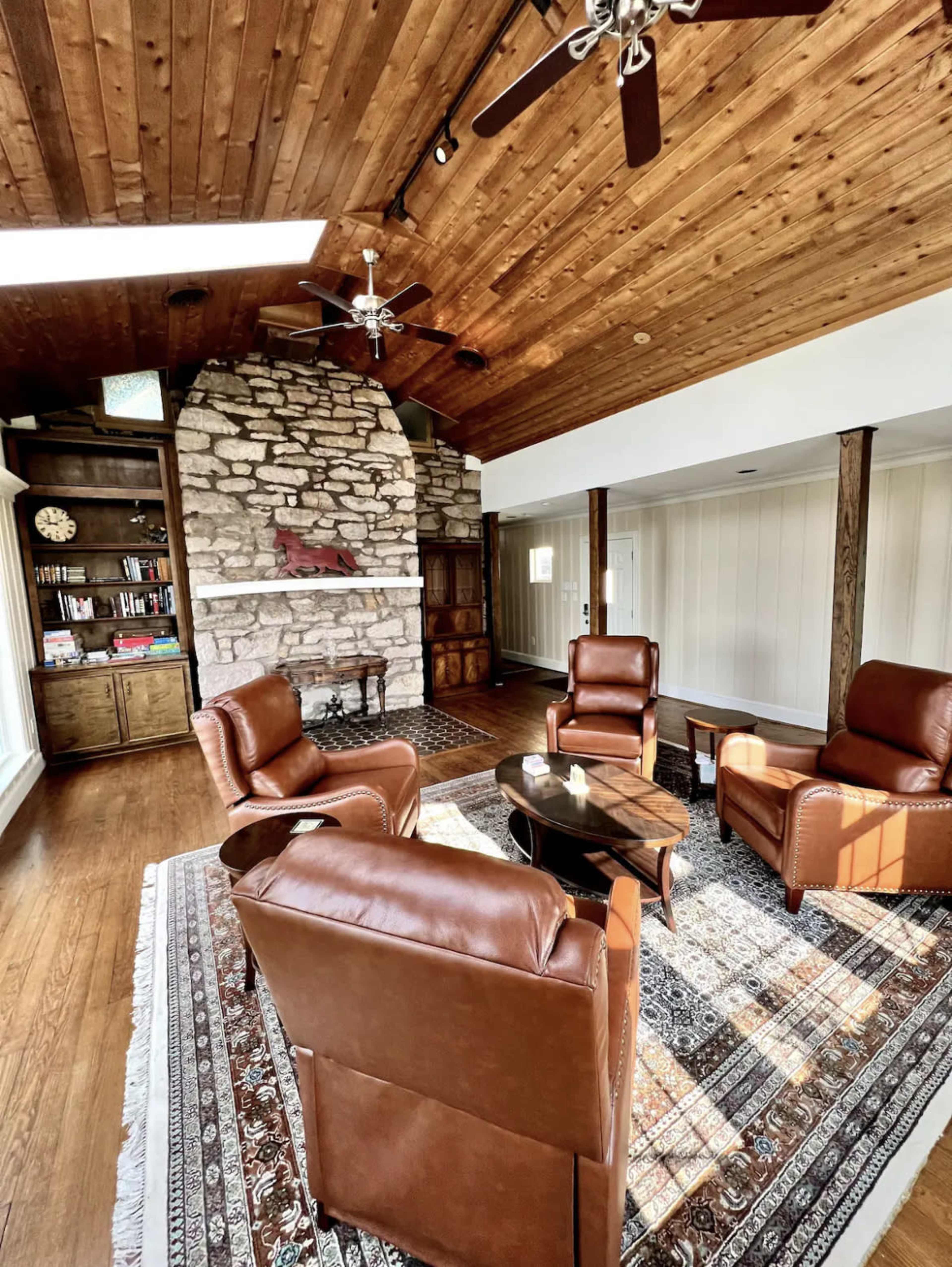 A cozy living room featuring a stone fireplace, wooden ceiling, and four brown leather chairs arranged around a circular table on a patterned rug.