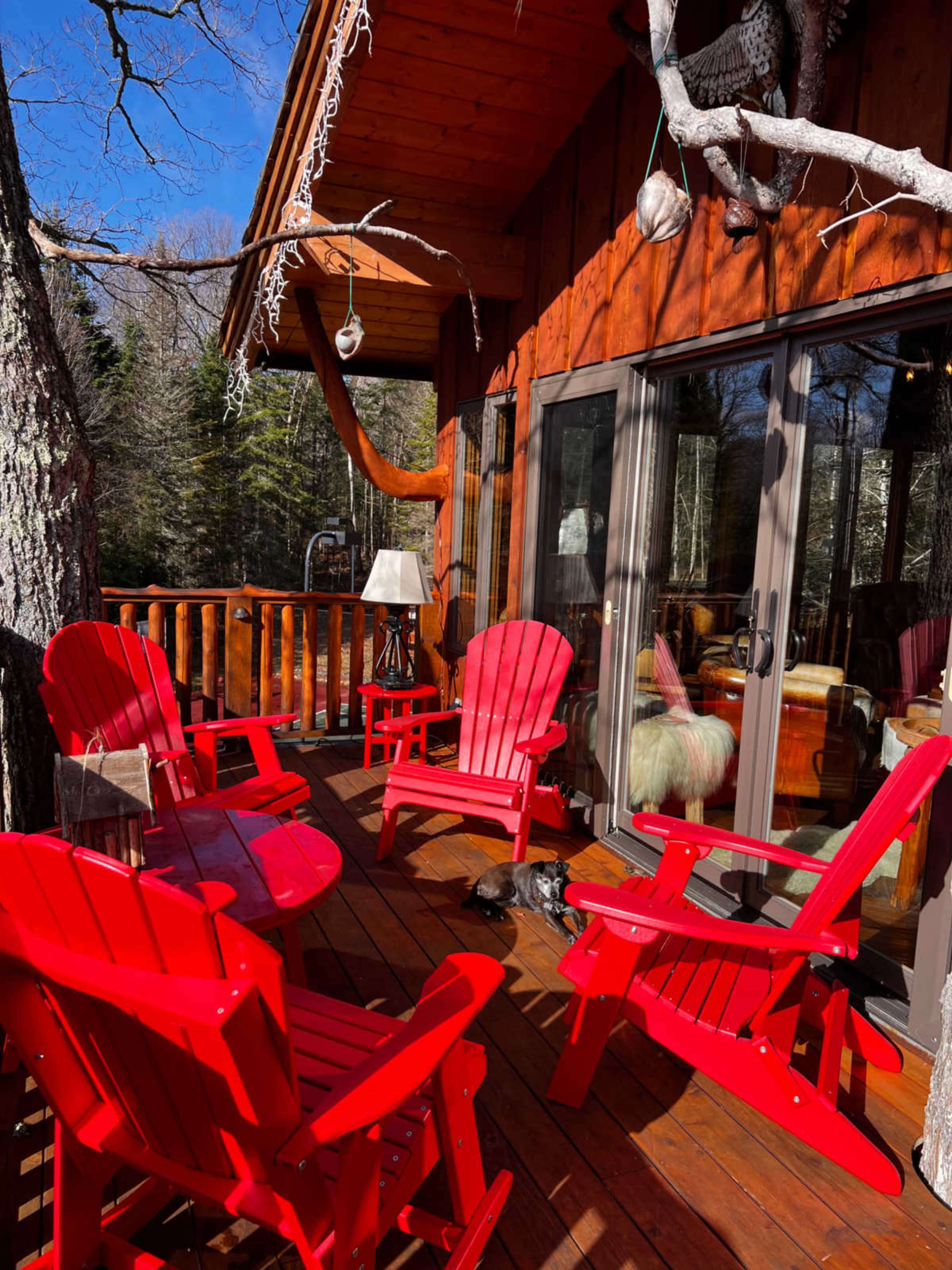 A wooden deck with red Adirondack chairs arranged around a small table, beside a large window with a view of a forest.