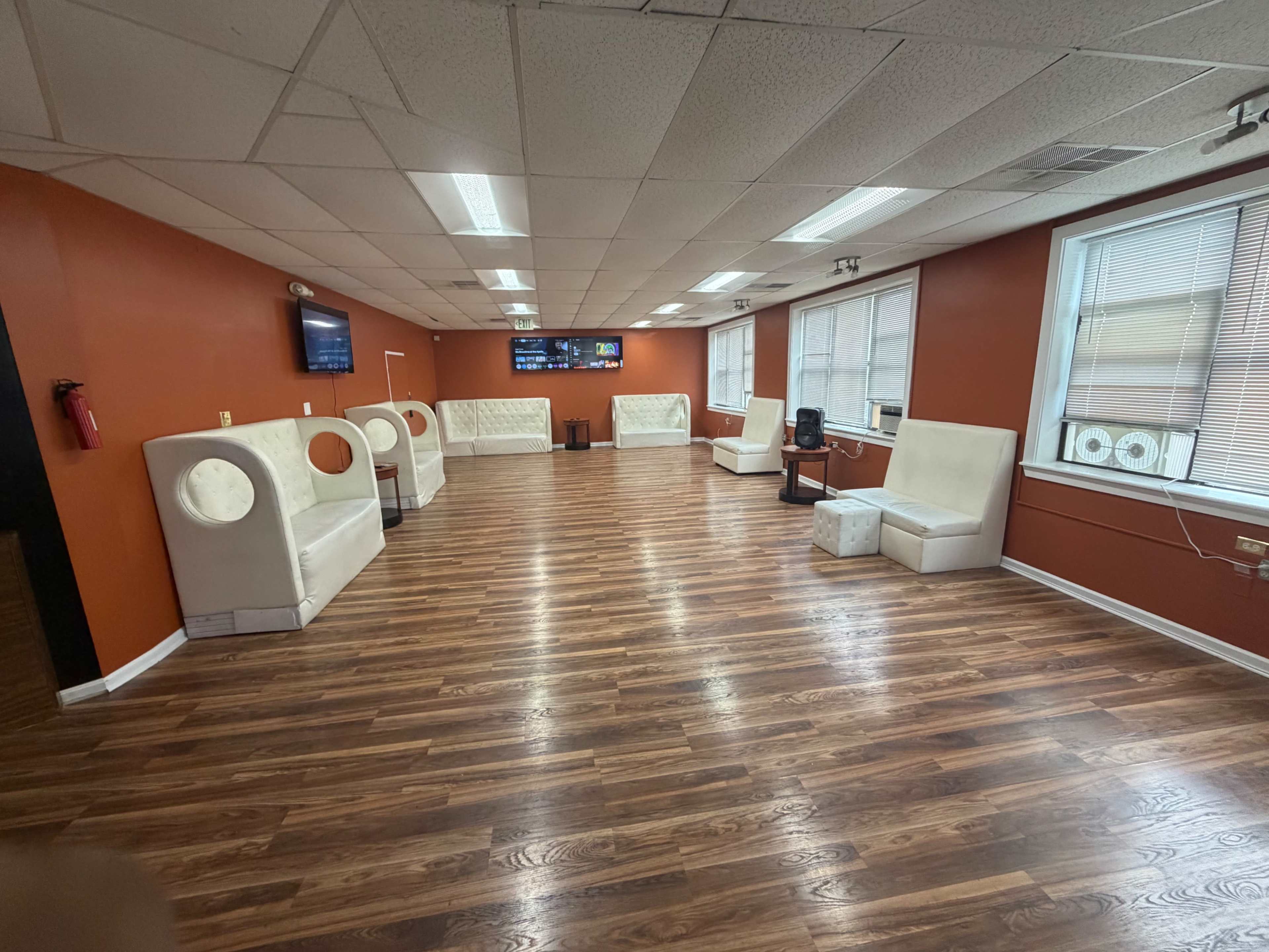 The image shows a spacious room with a hardwood floor and orange-painted walls, featuring white seating arranged around the perimeter.
