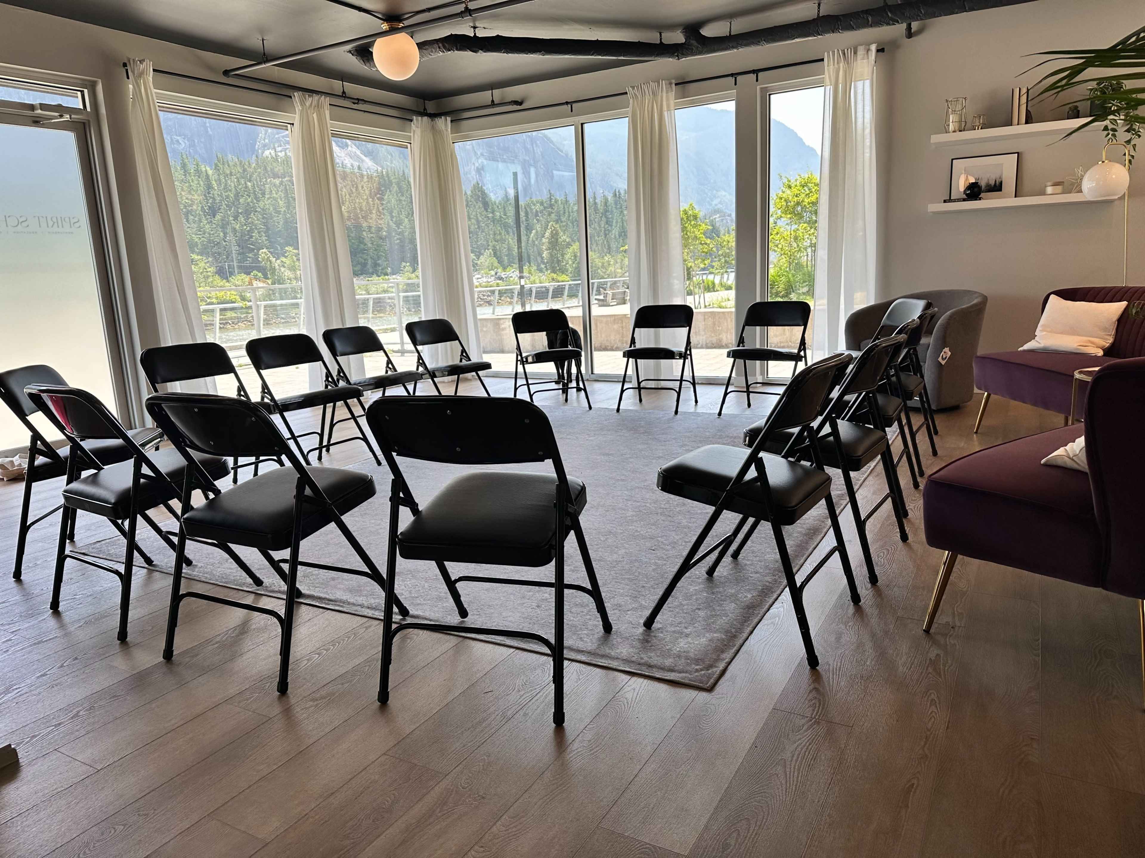 A room with large windows overlooks a mountain view and features a circle of black folding chairs arranged on a rug.