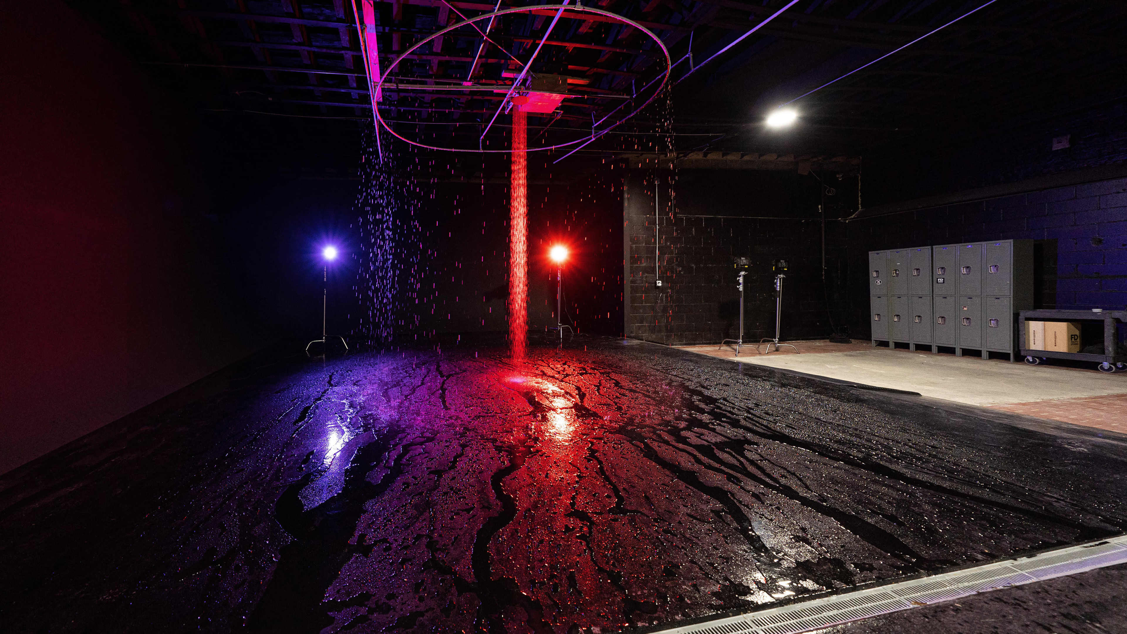 A dimly lit space with a circular structure hanging from the ceiling, water cascading down onto a shiny, wet floor, alongside colored lights creating an atmospheric effect.