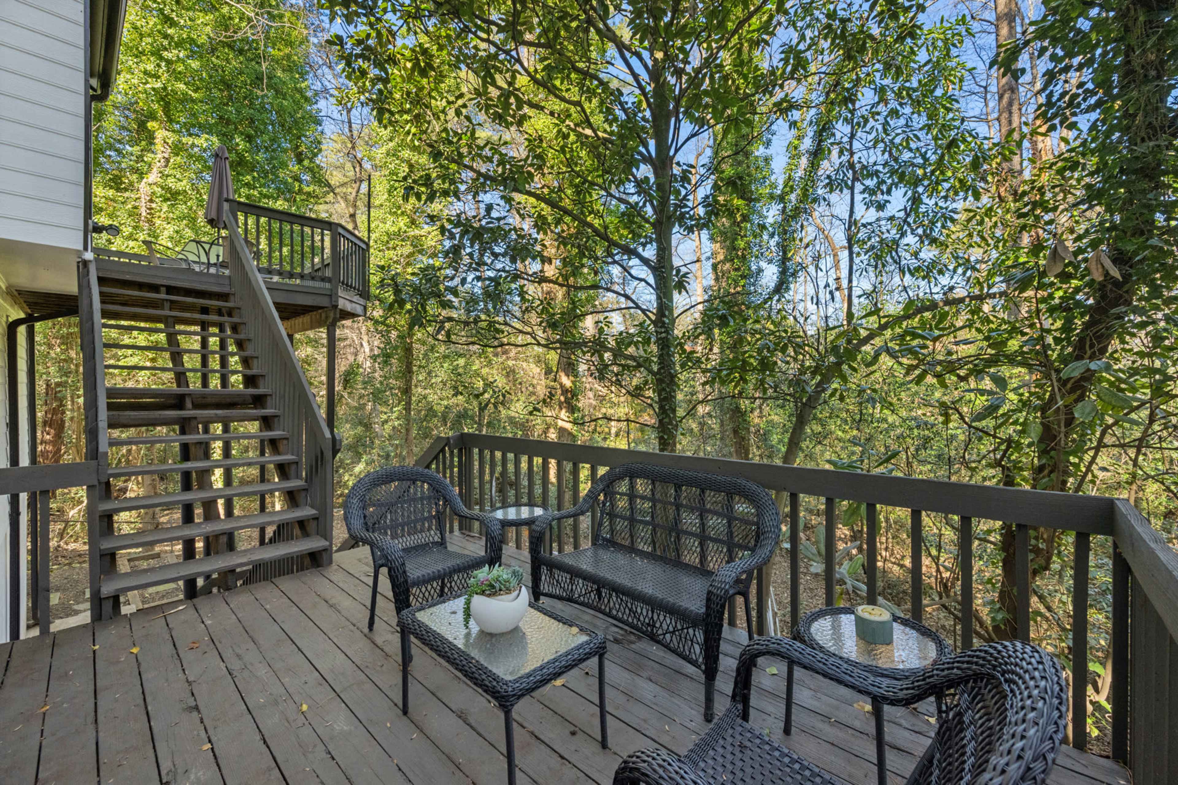 The image shows a wooden deck with black wicker furniture, surrounded by tall trees and greenery.