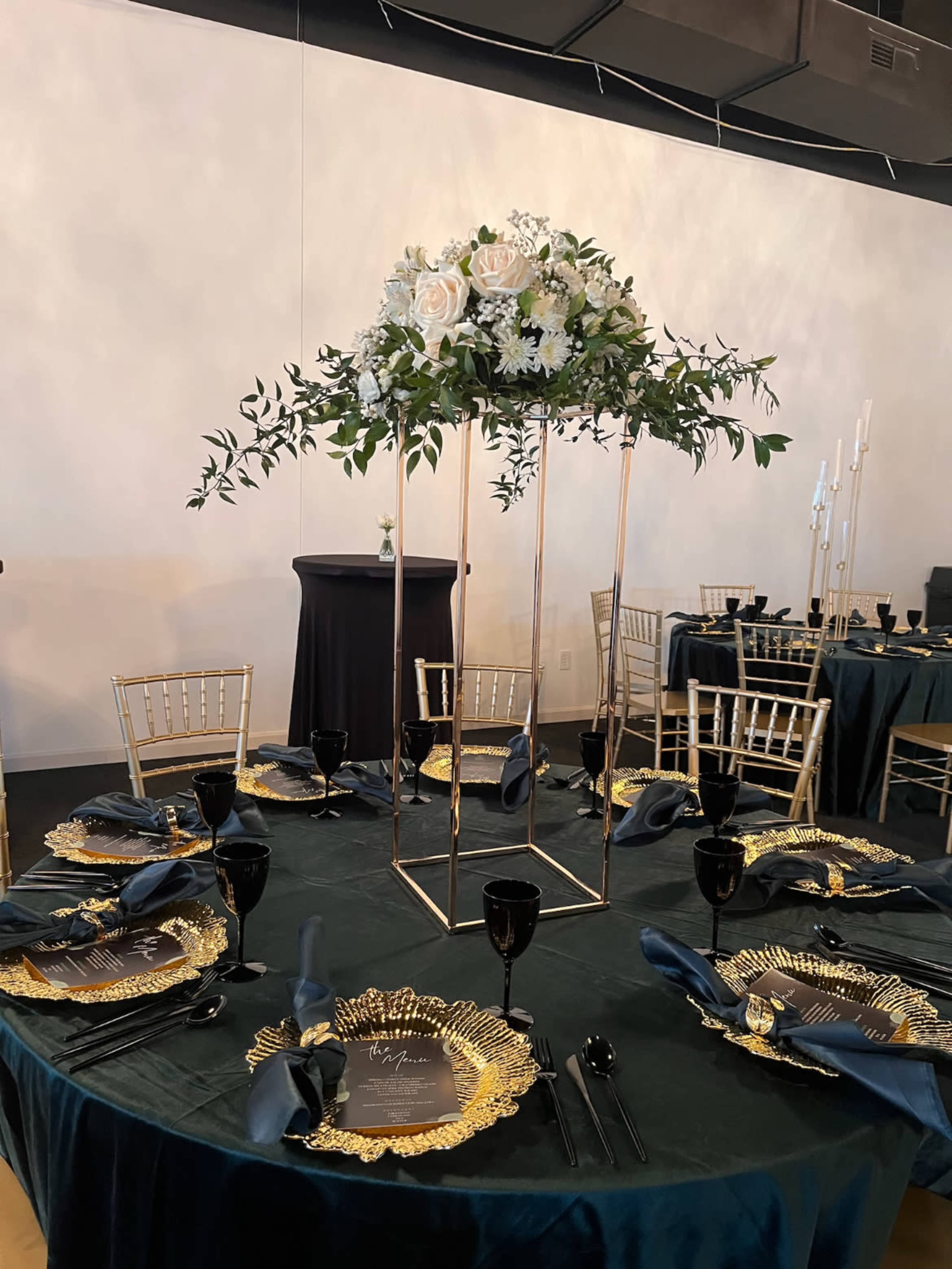 A formal dining table is set with dark green cloth, gold-rimmed plates, and black glasses, featuring a tall floral centerpiece with white roses.