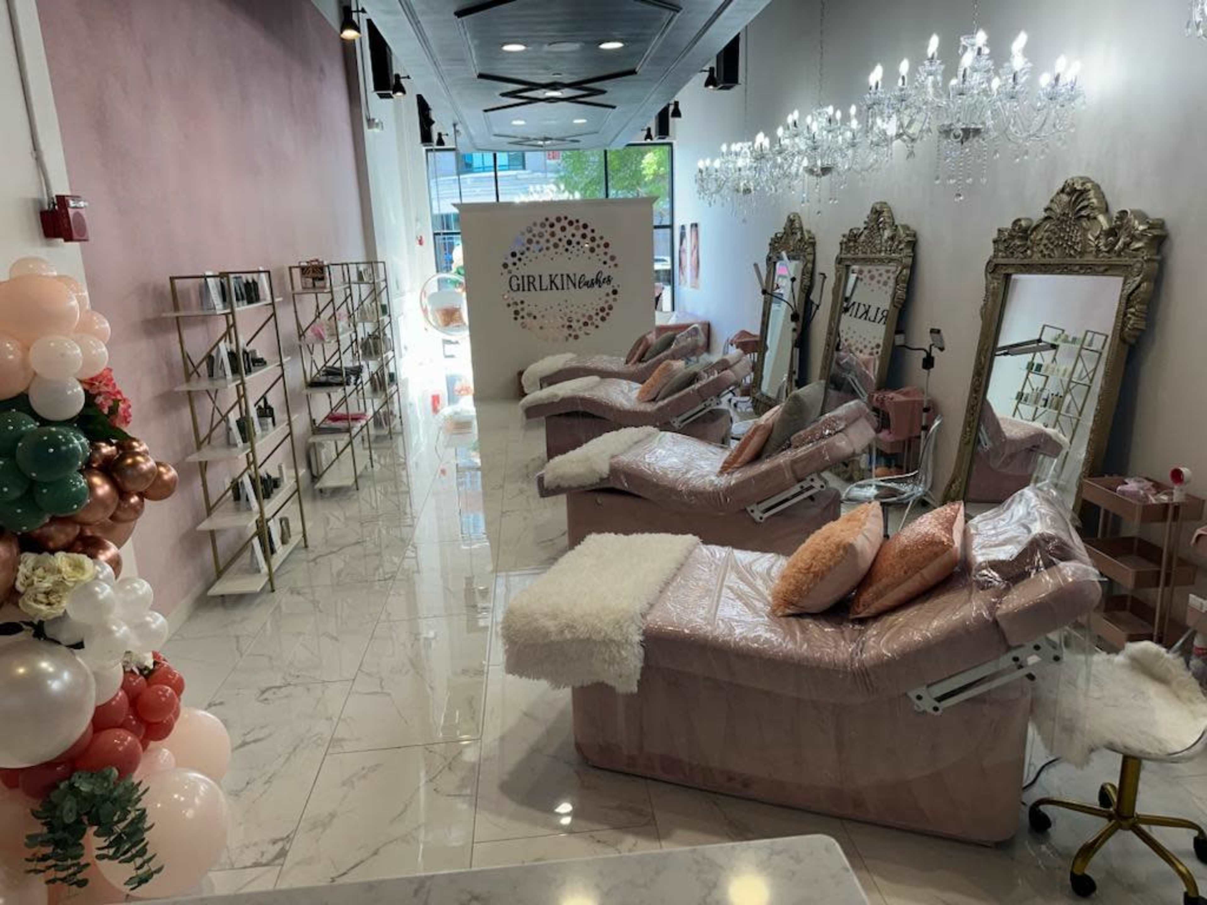 The image shows a modern beauty salon with pink treatment beds, ornate mirrors, and chandeliers, featuring decorative balloons along one side.