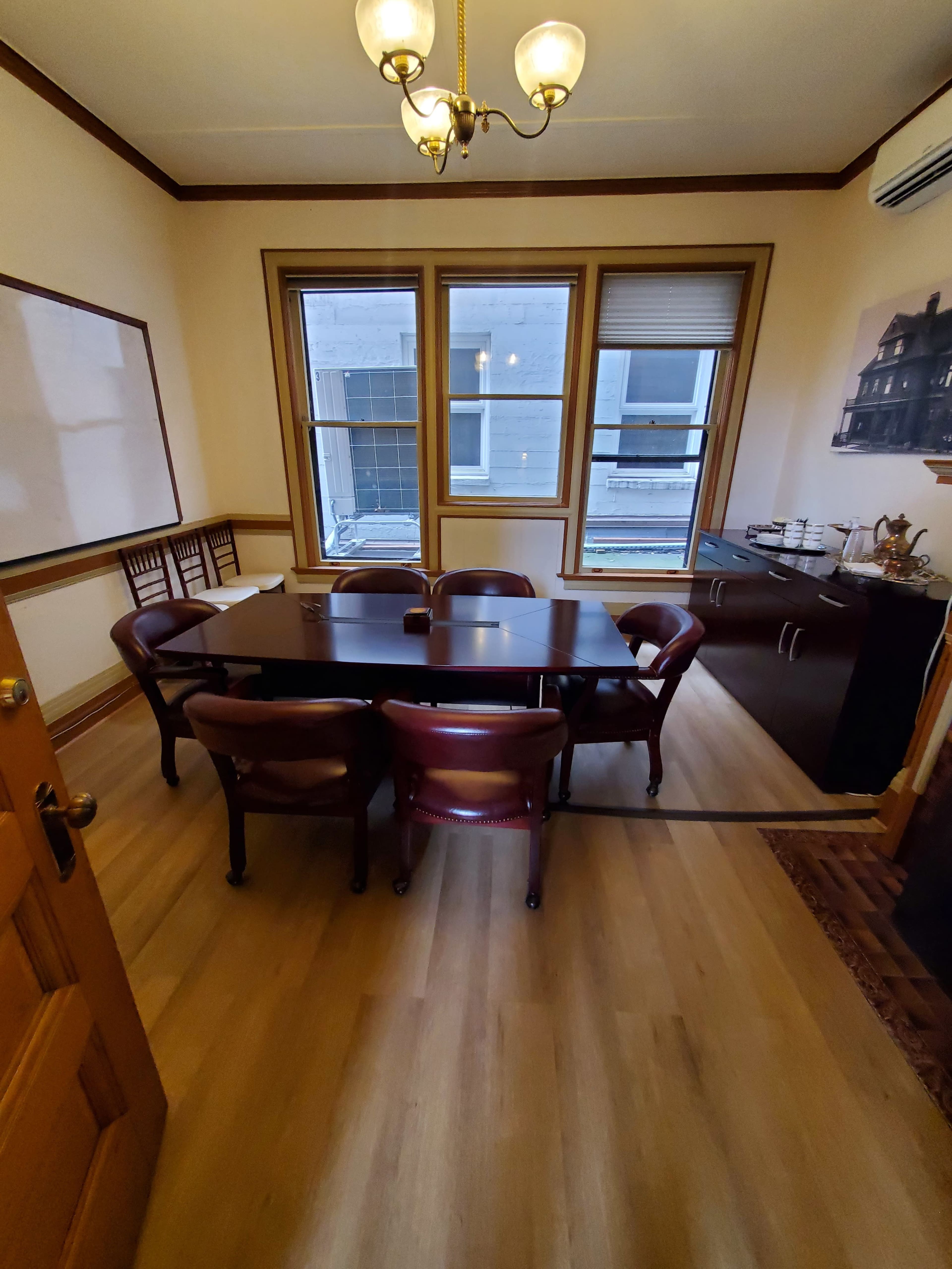 A small conference room with a dark wooden table surrounded by six chairs, large windows, and a side cabinet.