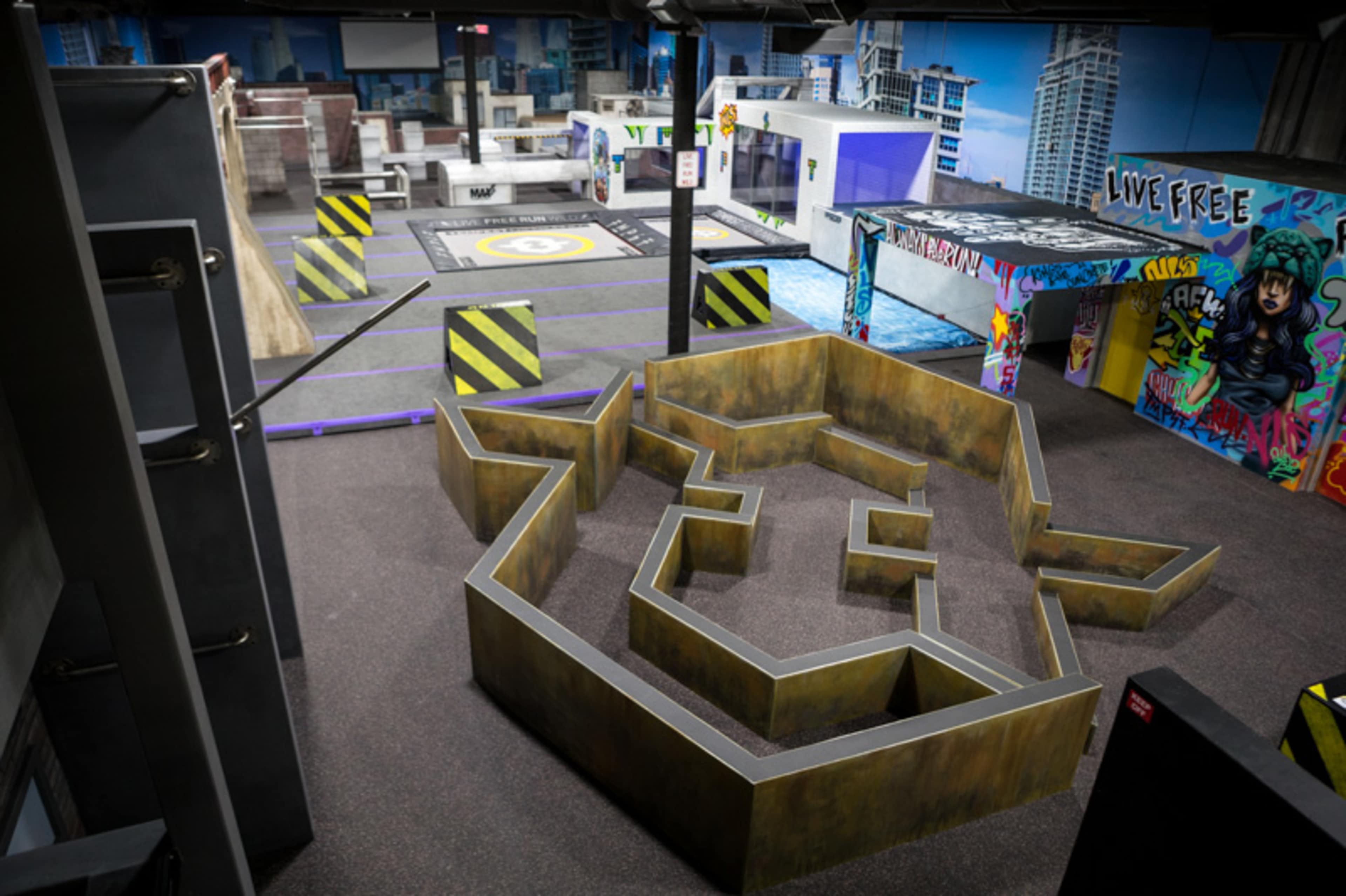 The image shows an indoor obstacle course with various structures, including a maze-like layout, colorful graffiti on the walls, and distinct zones marked by black and yellow barriers.