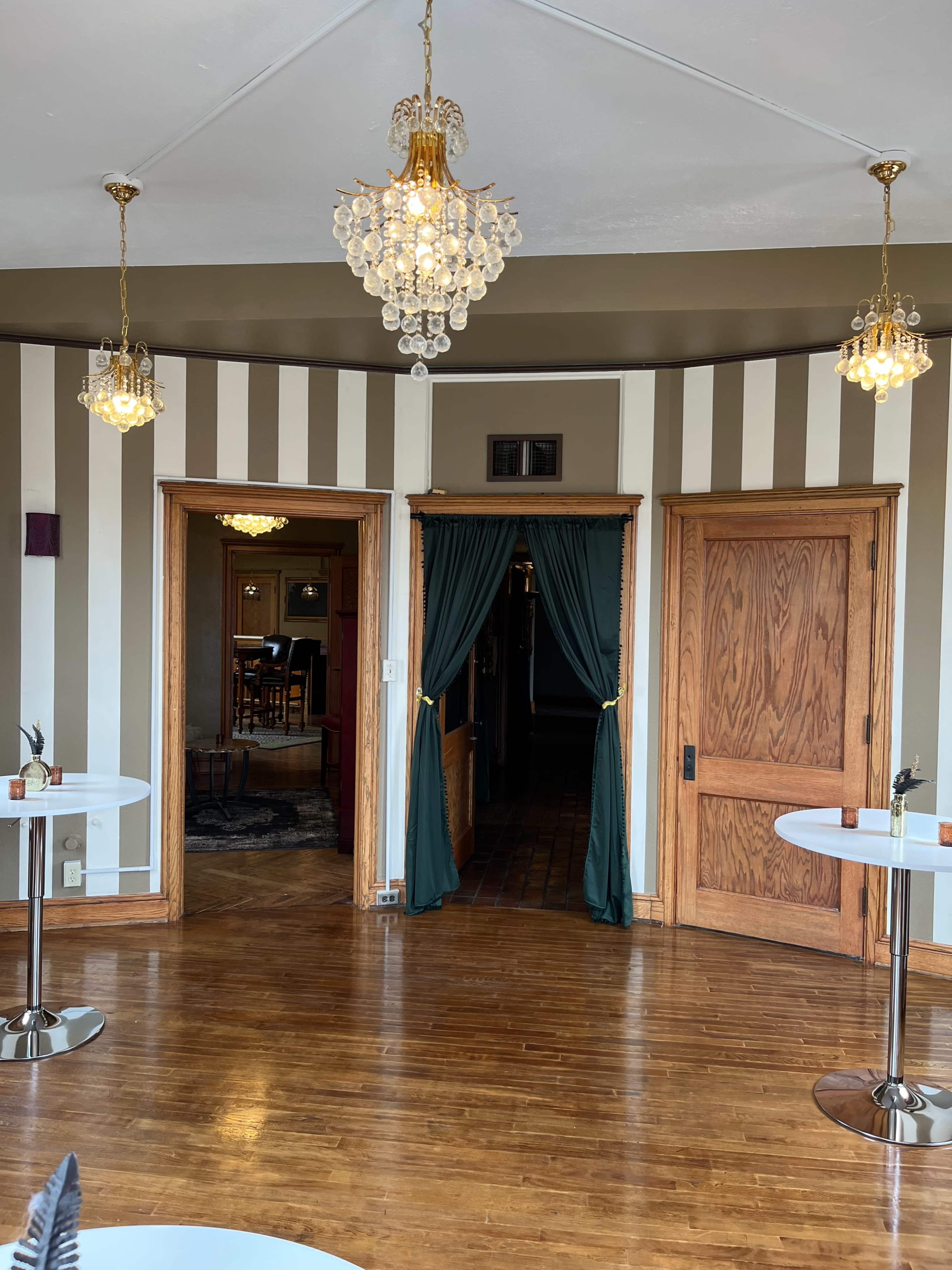 A room with wooden floors, striped walls, and two entrances, each adorned with curtains, beneath hanging chandeliers.