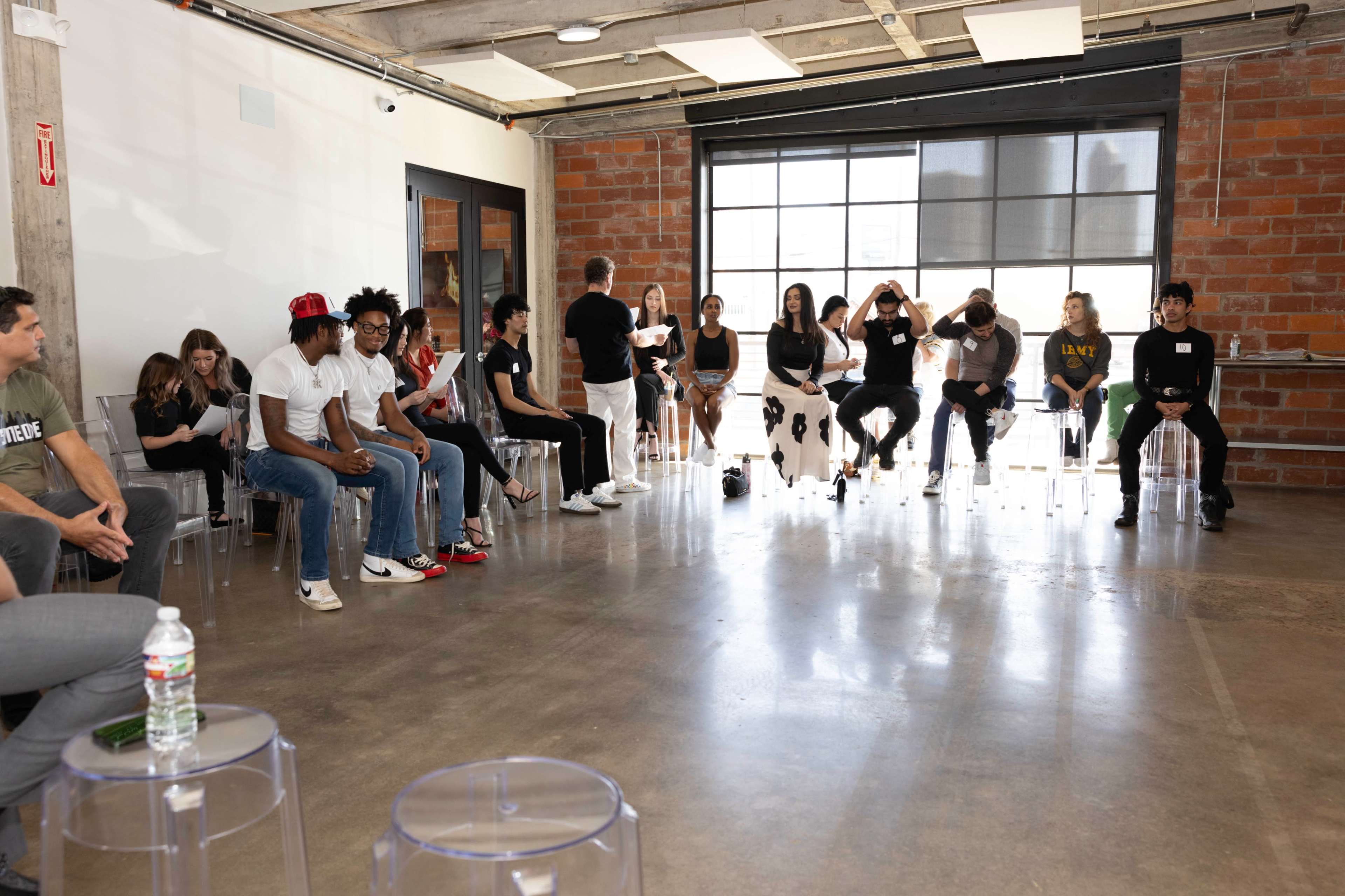 A group of young adults sits in a modern room with a concrete floor and large windows, engaged in conversation.