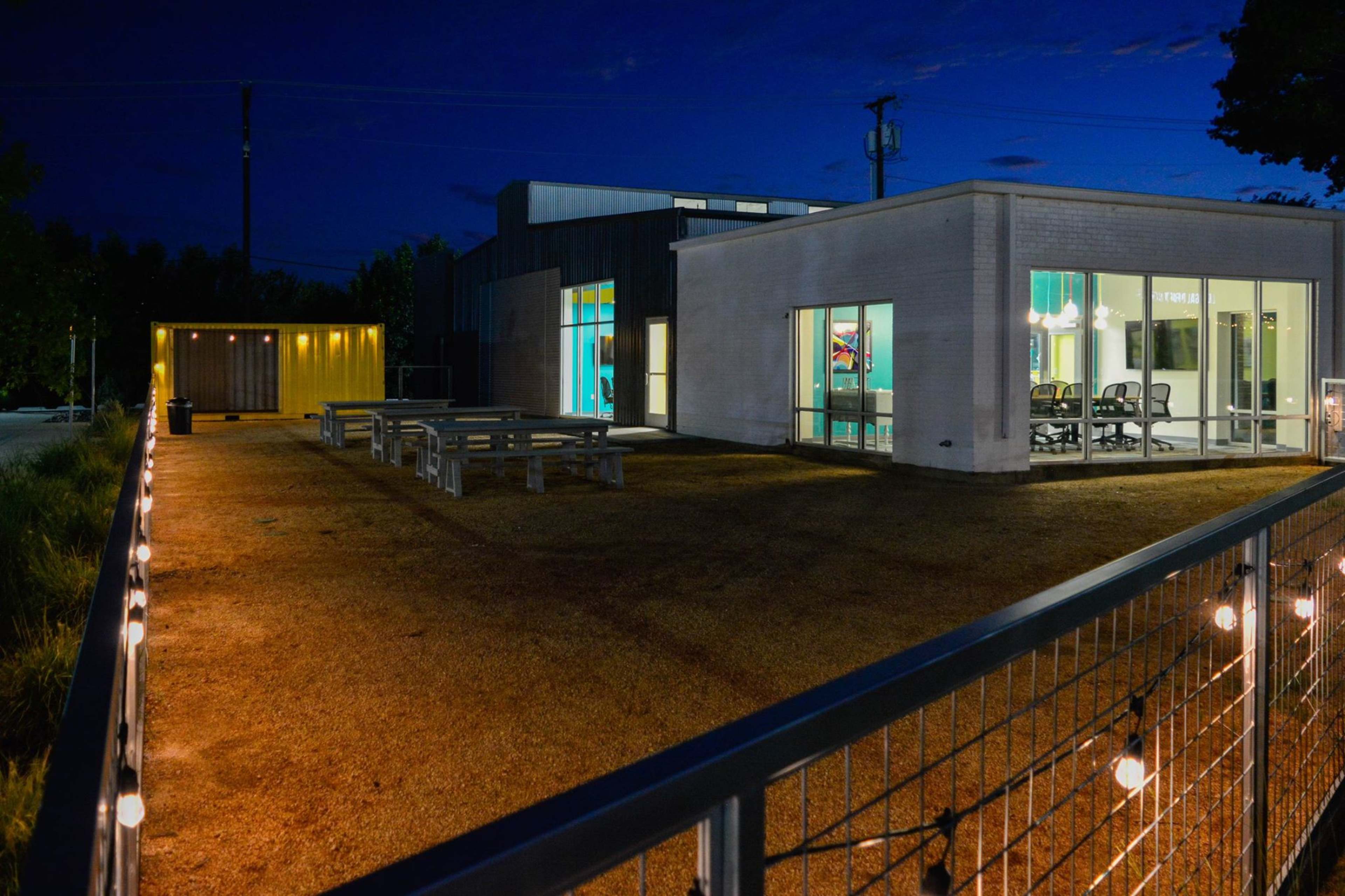 The photo shows a modern building with large windows and outdoor seating illuminated by string lights during twilight.