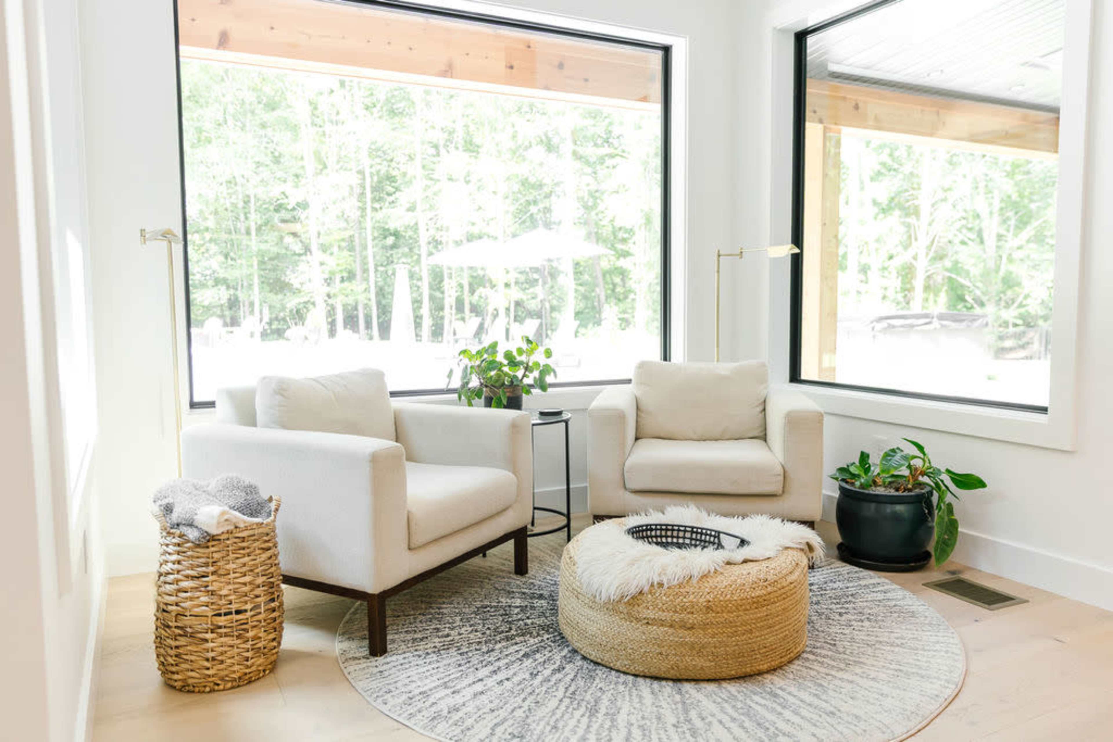The image shows a modern sitting area with two light-colored armchairs, a round woven pouf, and a small table, surrounded by large windows that allow natural light to enter.