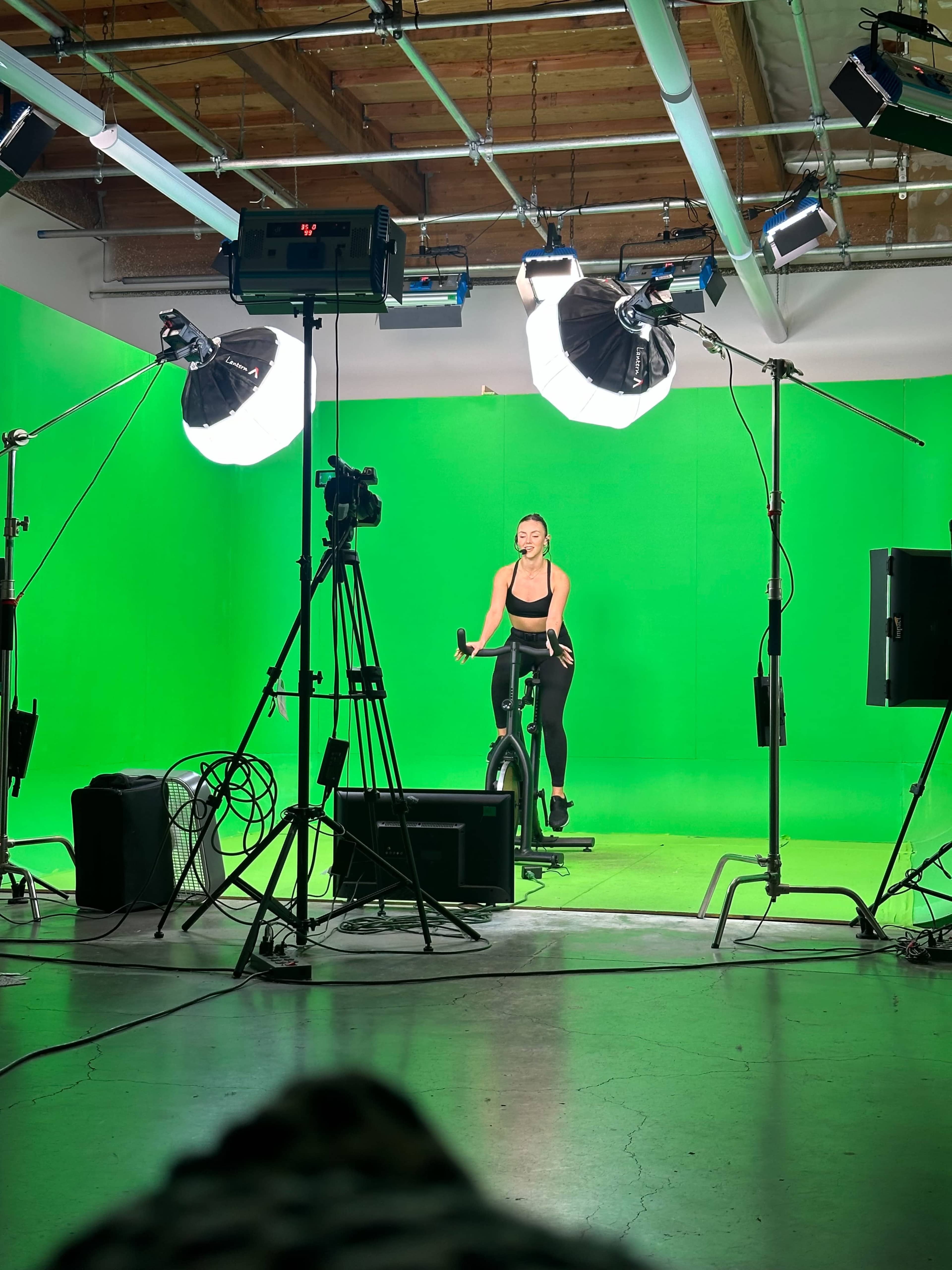 A person is riding an exercise bike in front of a lit green screen, surrounded by cameras and studio lighting equipment.